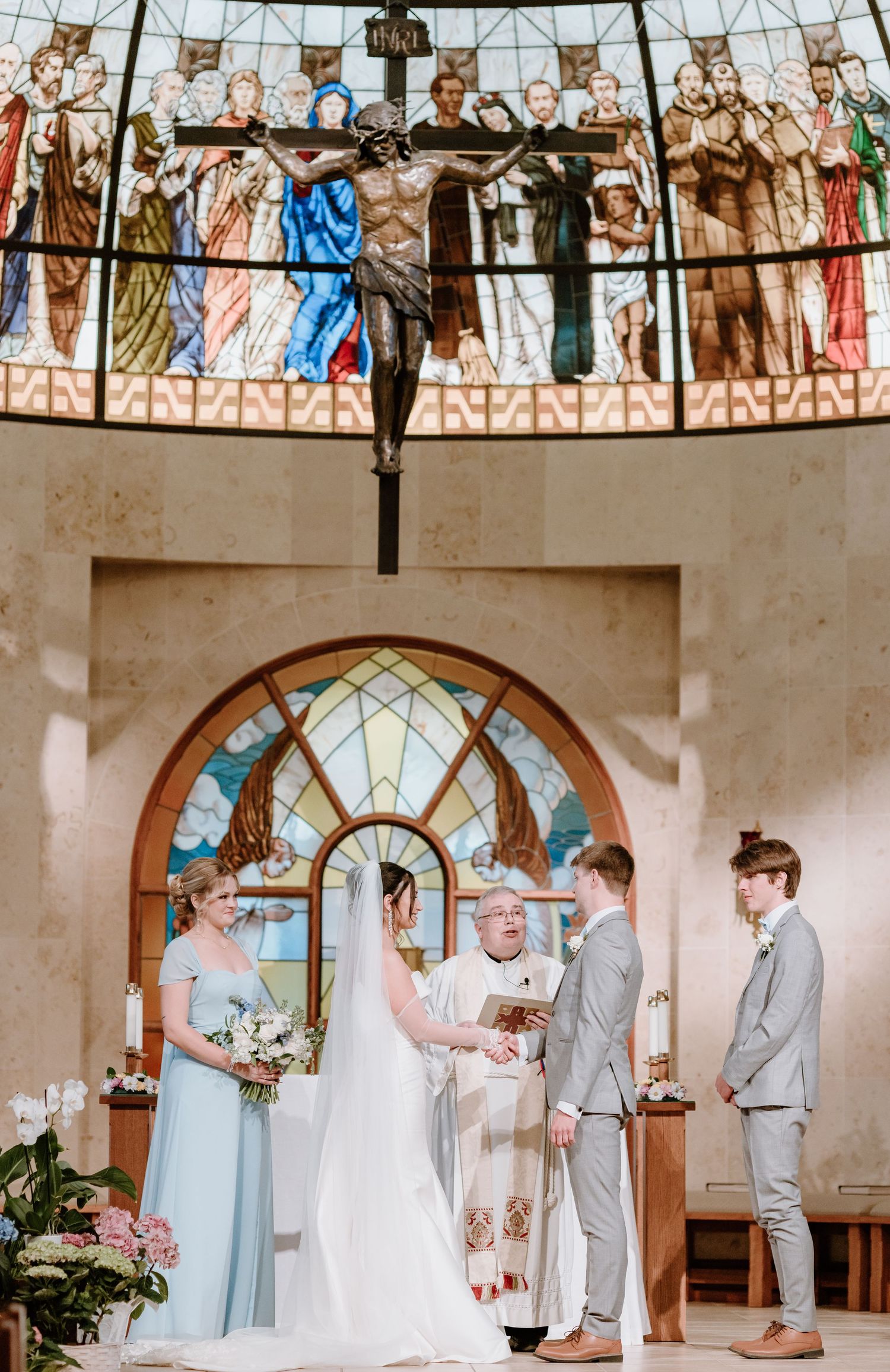 A wedding ceremony takes place in a chapel with stained glass windows and religious artwork on the walls.