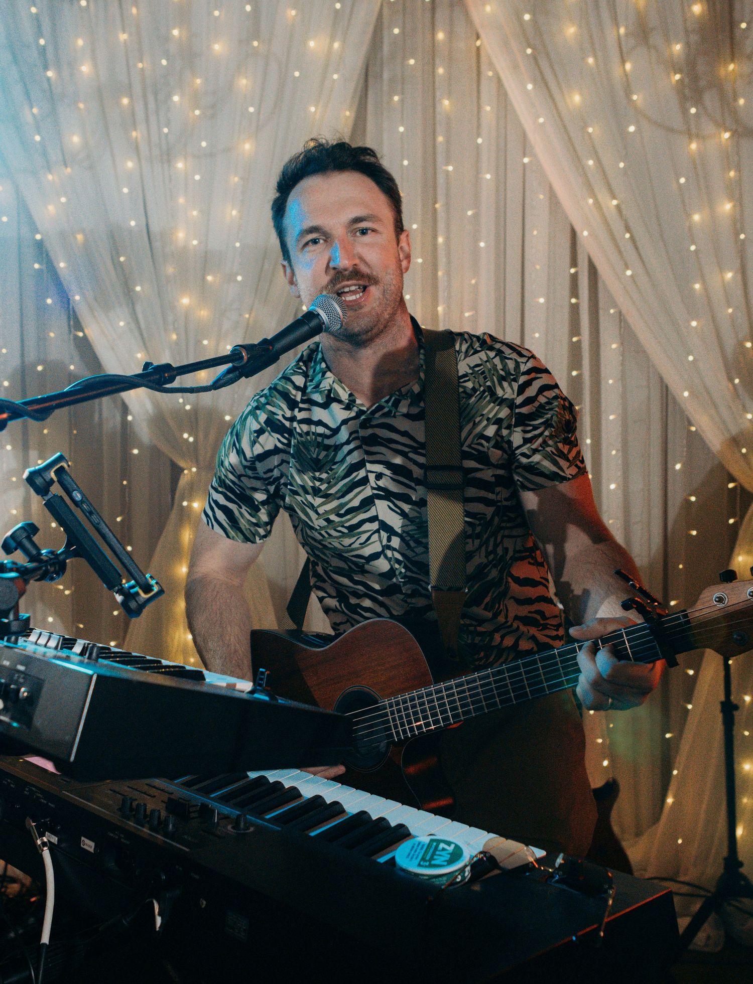 A musician performs with an acoustic guitar and keyboard against a backdrop of twinkling fairy lights.