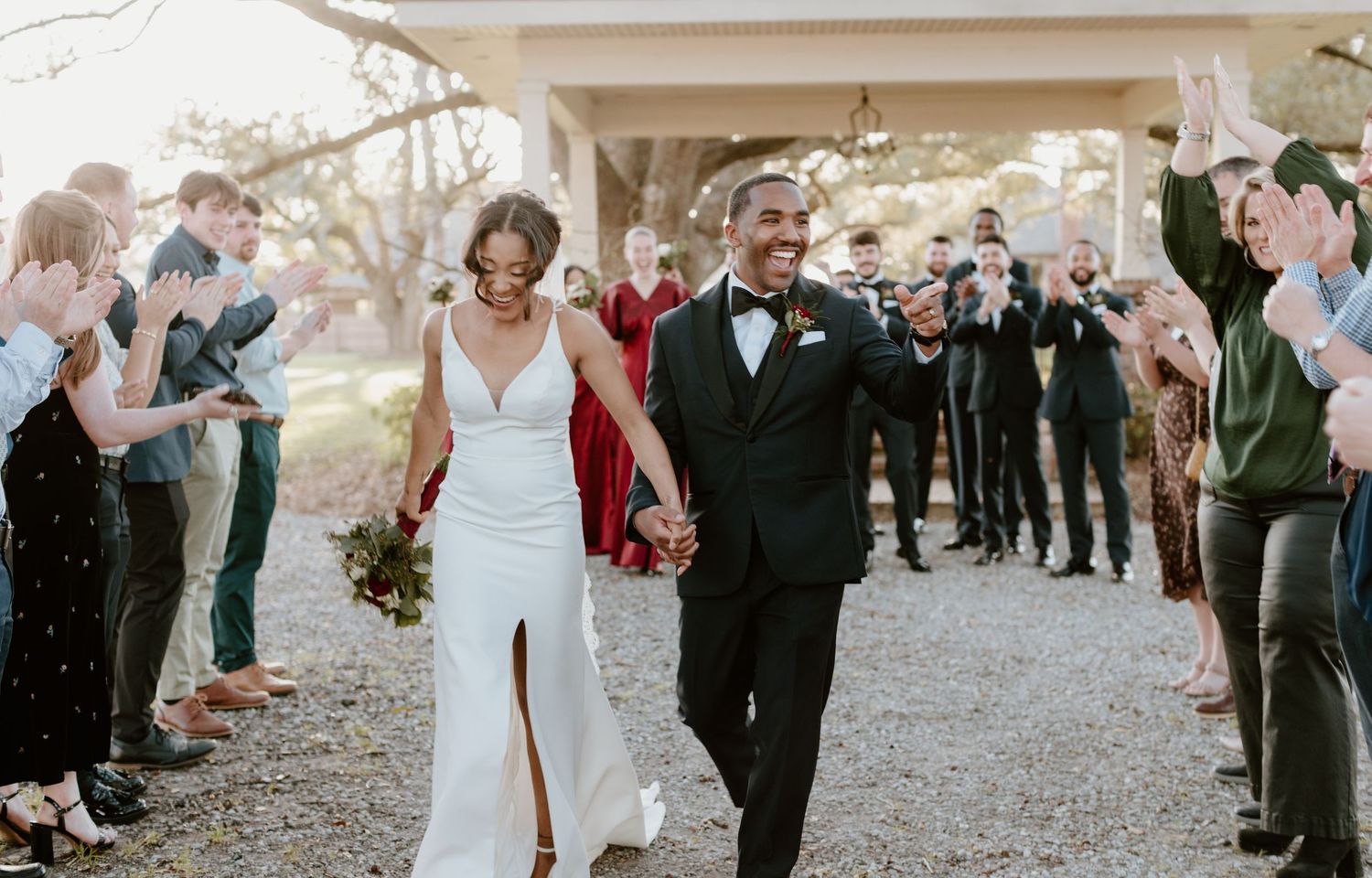 Joyful newlyweds walk down outdoor path as guests clap and cheer during wedding ceremony exit.