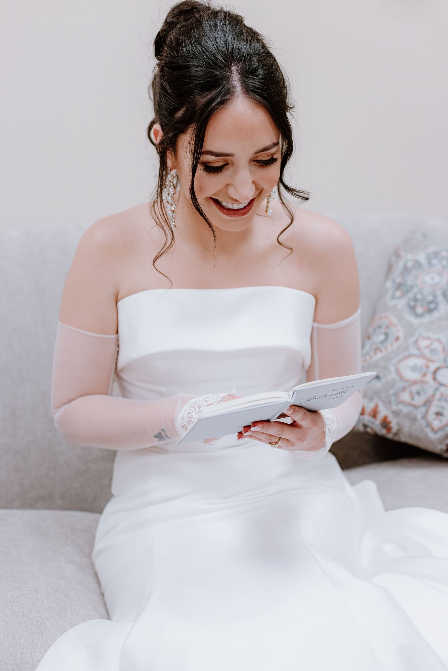 A bride in a white wedding dress reads a letter while sitting on a couch, wearing delicate sheer sleeves.