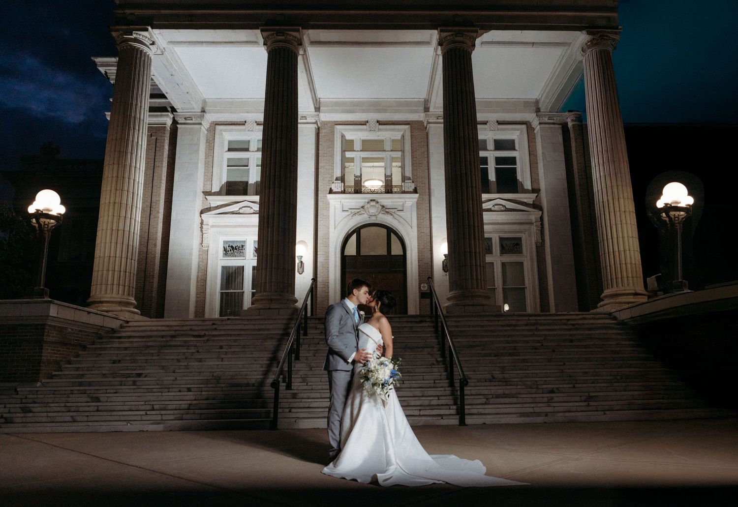 A couple embraces on the steps of a grand historic building with dramatic columns and lighting at night.