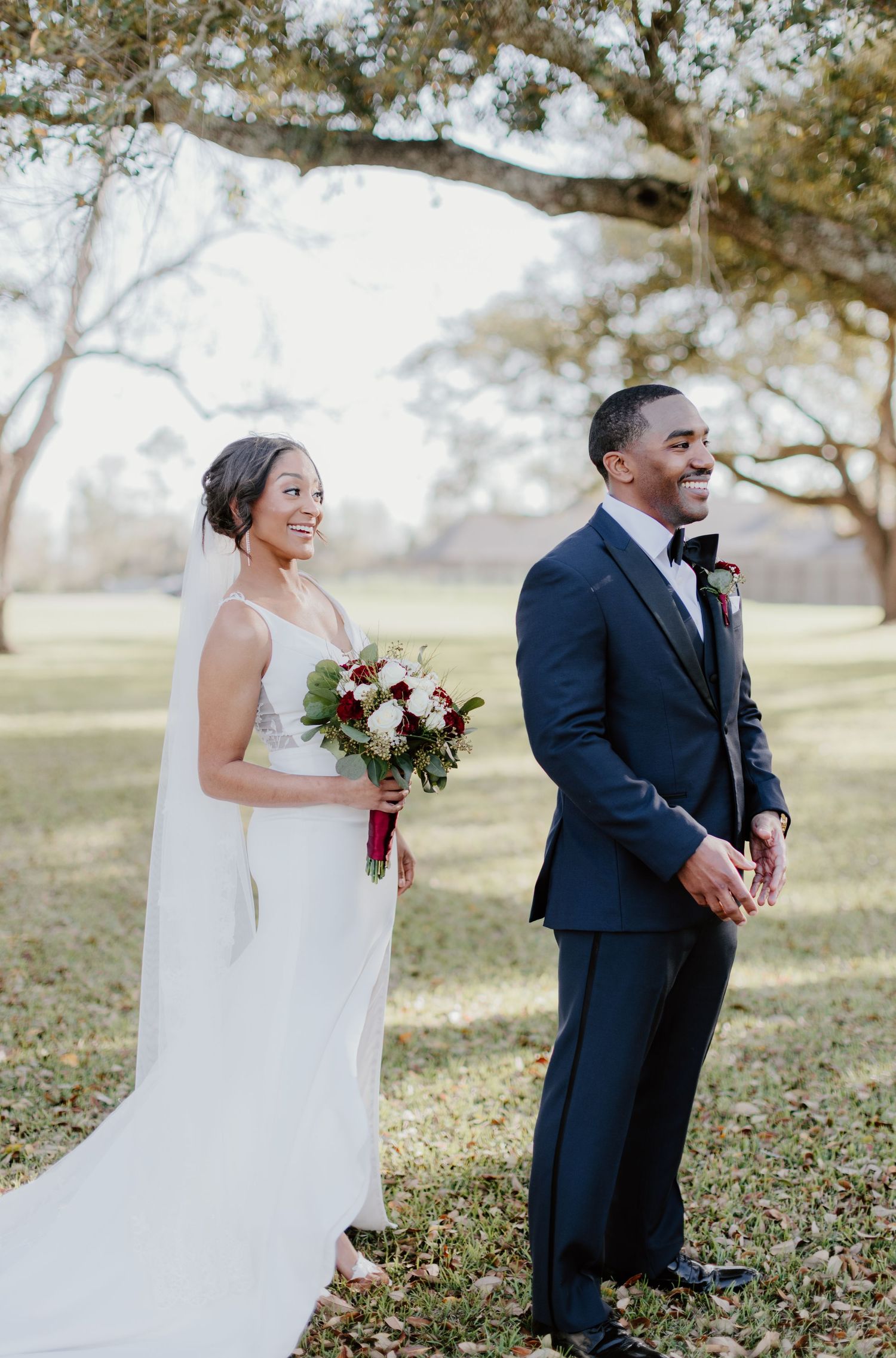 A newlywed couple stands in a dreamy outdoor setting with trees, sharing a joyful moment in formal wedding attire.