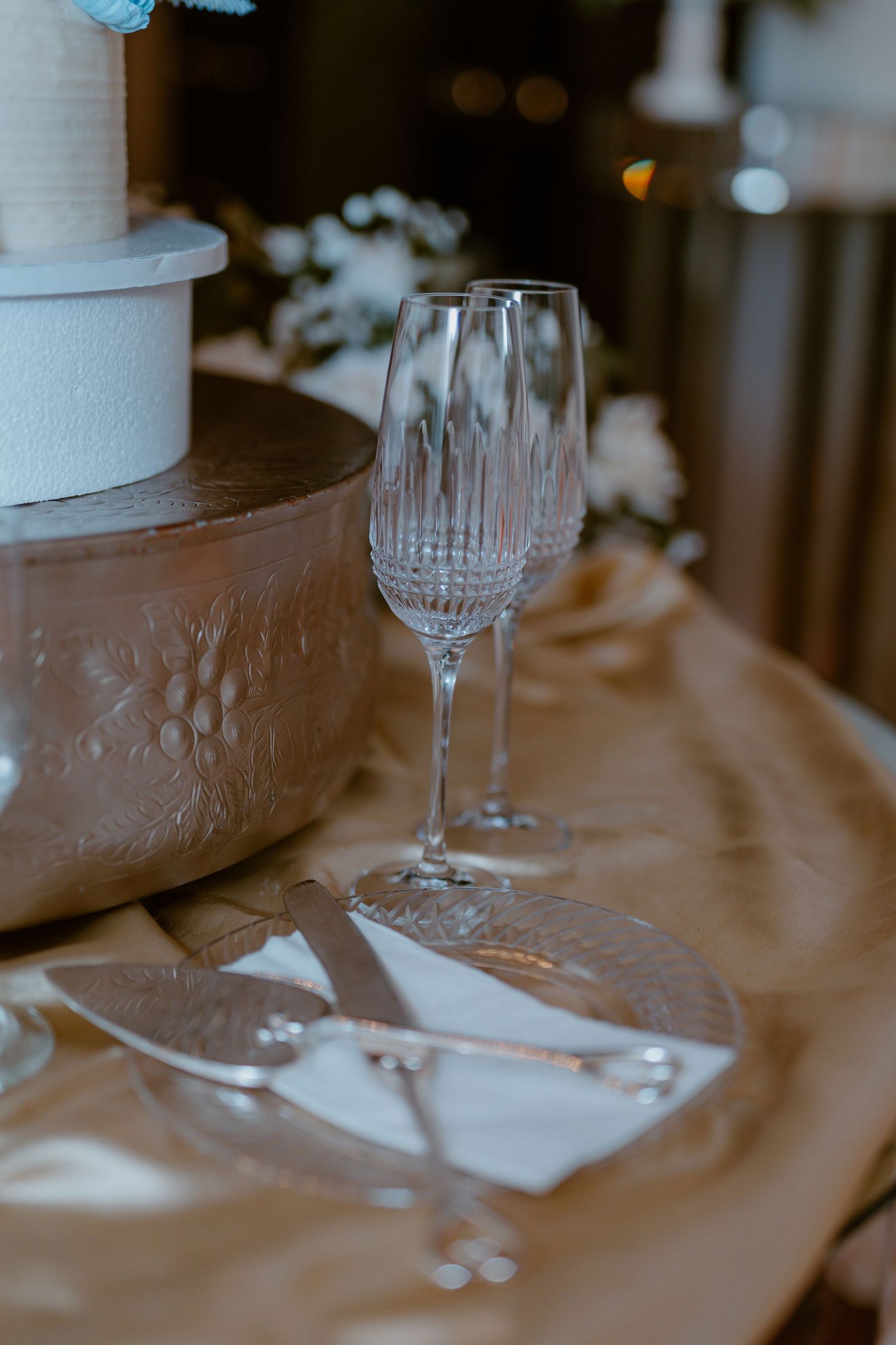 A crystal champagne flute and cake serving utensils rest on a satin tablecloth beside a wedding cake.