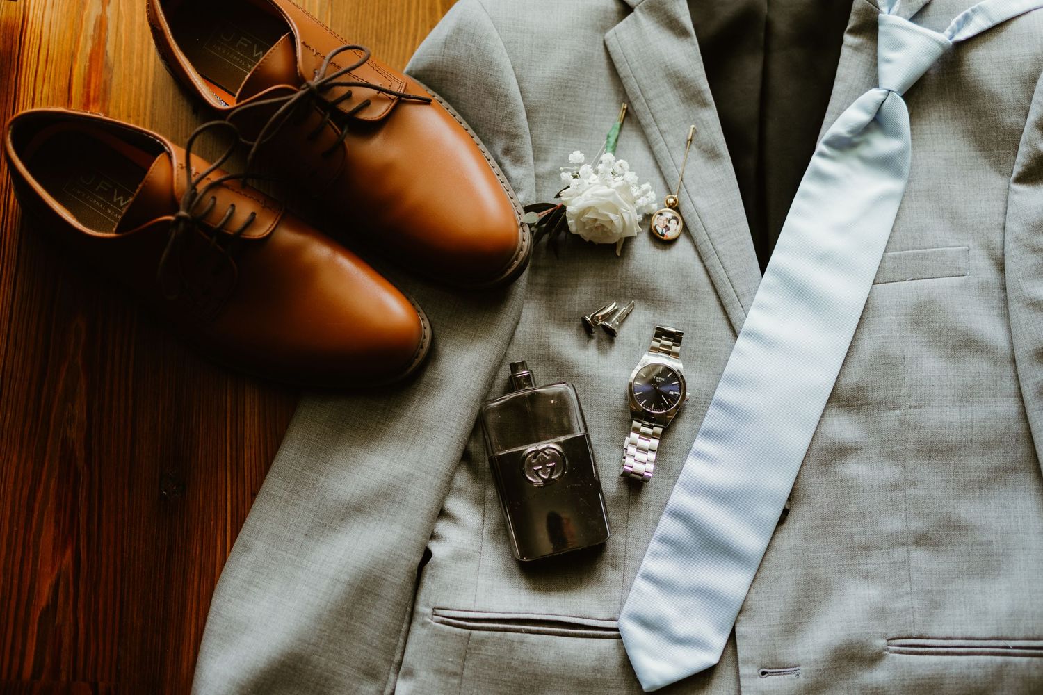 Elegant wedding accessories including brown leather shoes, gray suit jacket, watches and tie laid on wooden surface.