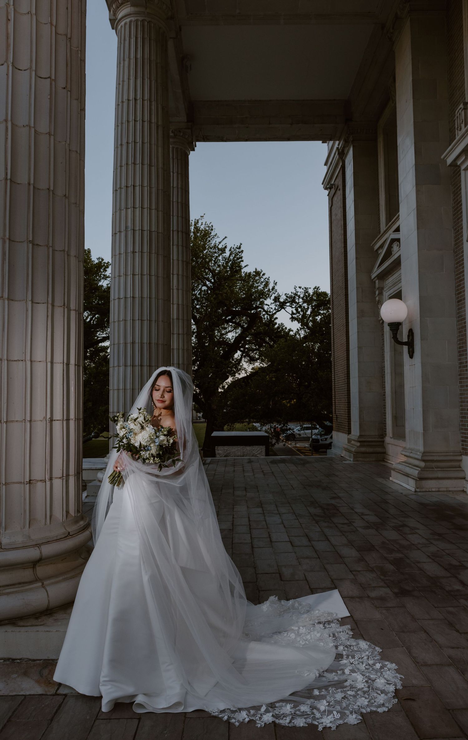 Elegant bridal portrait at dusk featuring a flowing white gown and veil against classic stone columns.