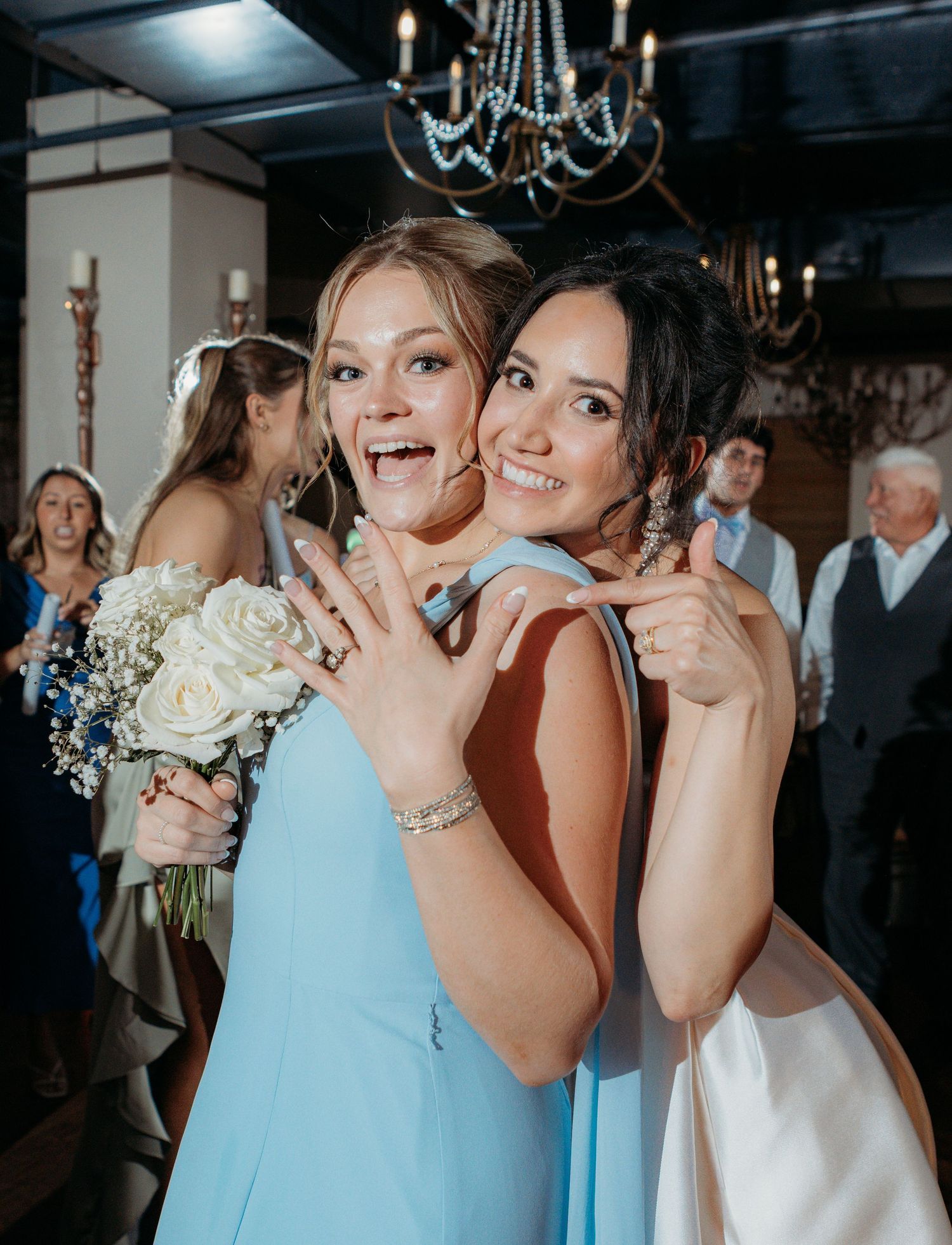 Two wedding attendees pose together in elegant dresses, showing off a ring while holding a white bouquet at a reception.