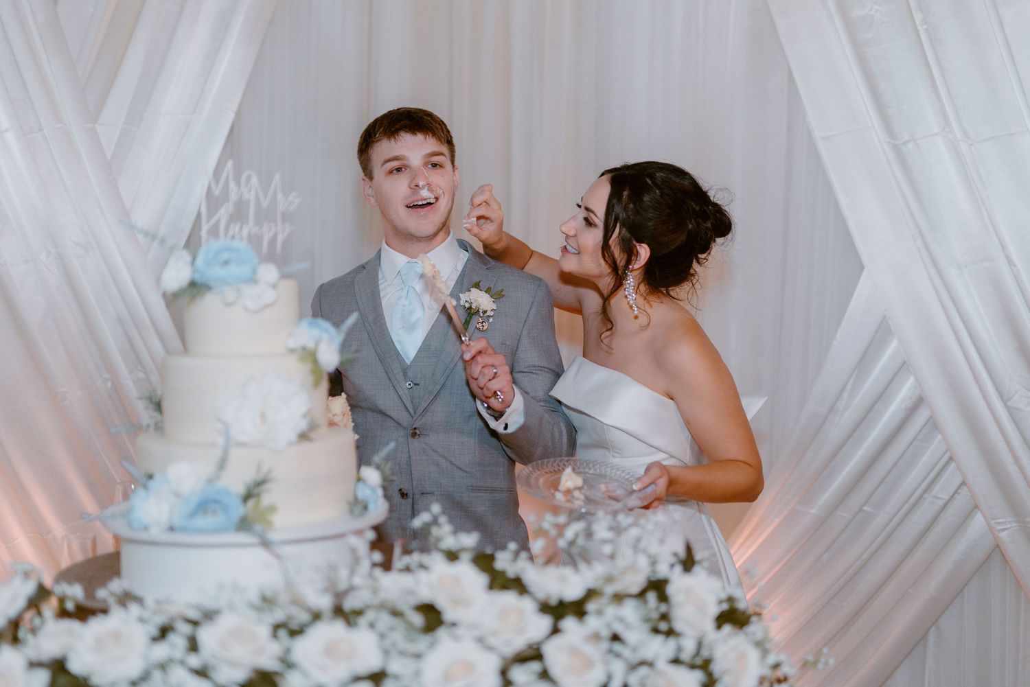 Couple at their wedding sharing a playful cake-tasting moment in front of elegant white drapery and tiered cake.