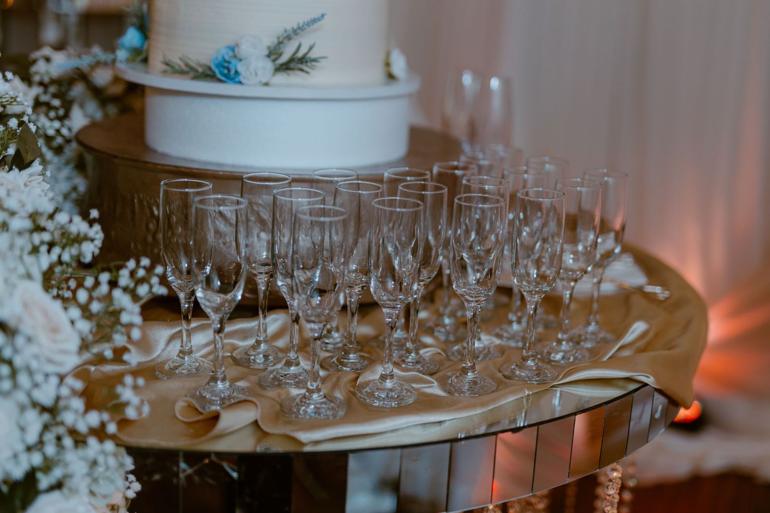 Empty champagne glasses arranged on a mirrored tray next to a white wedding cake with blue floral accents.