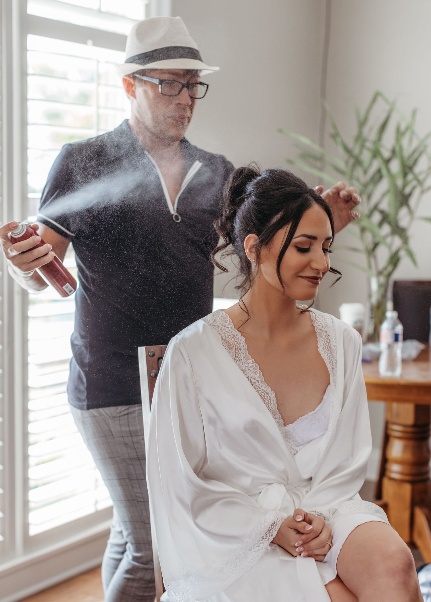 A hairstylist sprays hairspray while styling a client wearing a white silk robe in a sunlit room.