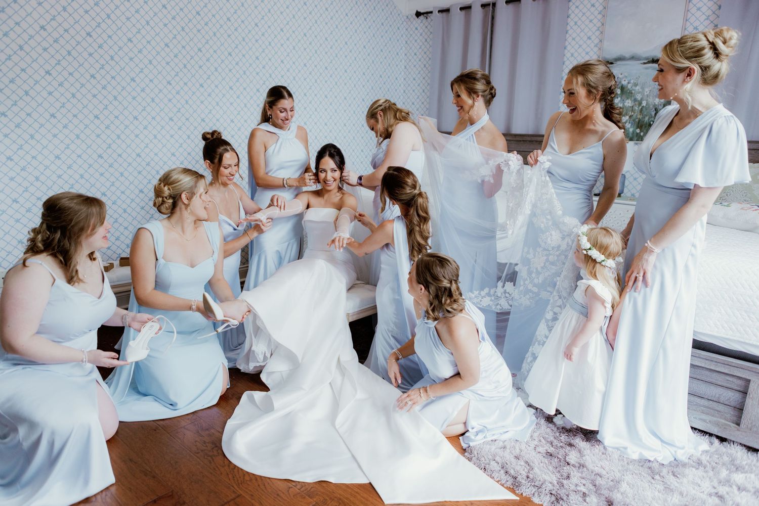 A group of bridesmaids in light blue dresses gather around a bride in a white gown during wedding preparations.