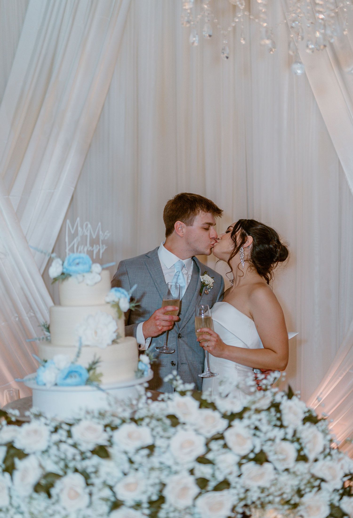 Newlyweds share a romantic kiss behind their elegant white wedding cake decorated with blue accents and white floral arrangements.