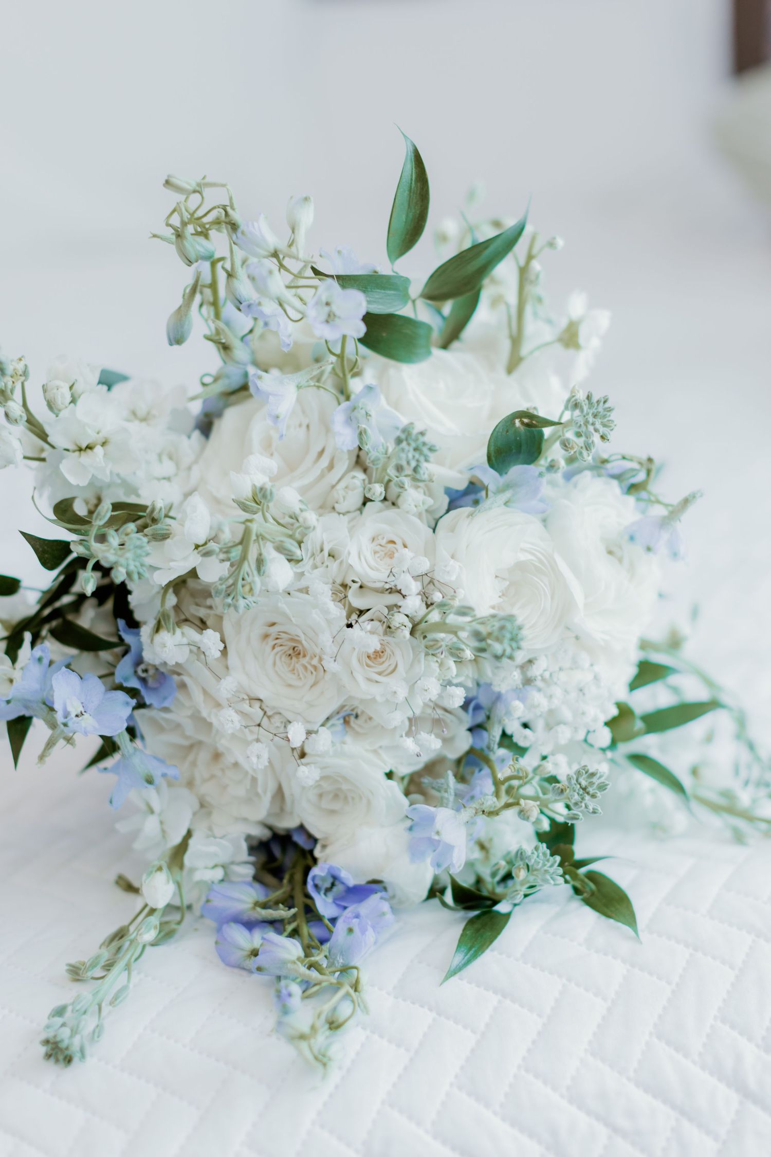 A delicate white and blue wedding bouquet with roses, greenery and pastel accents on a textured white surface.
