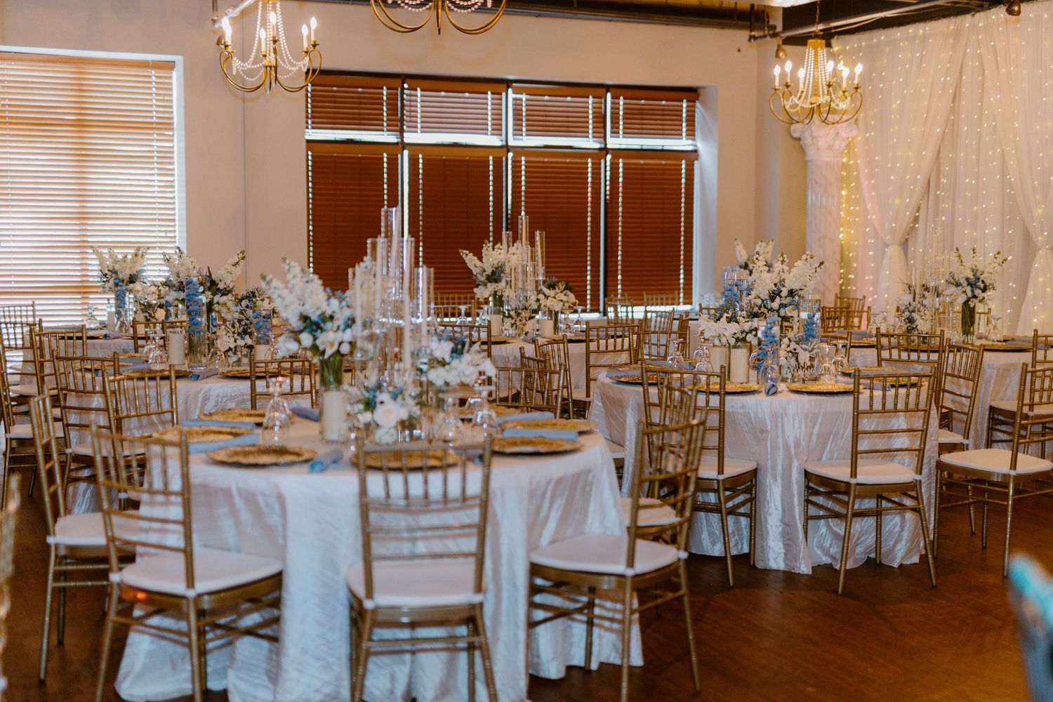 Elegant wedding reception venue with white linens, gold chiavari chairs, and crystal chandeliers in warm ambient lighting.