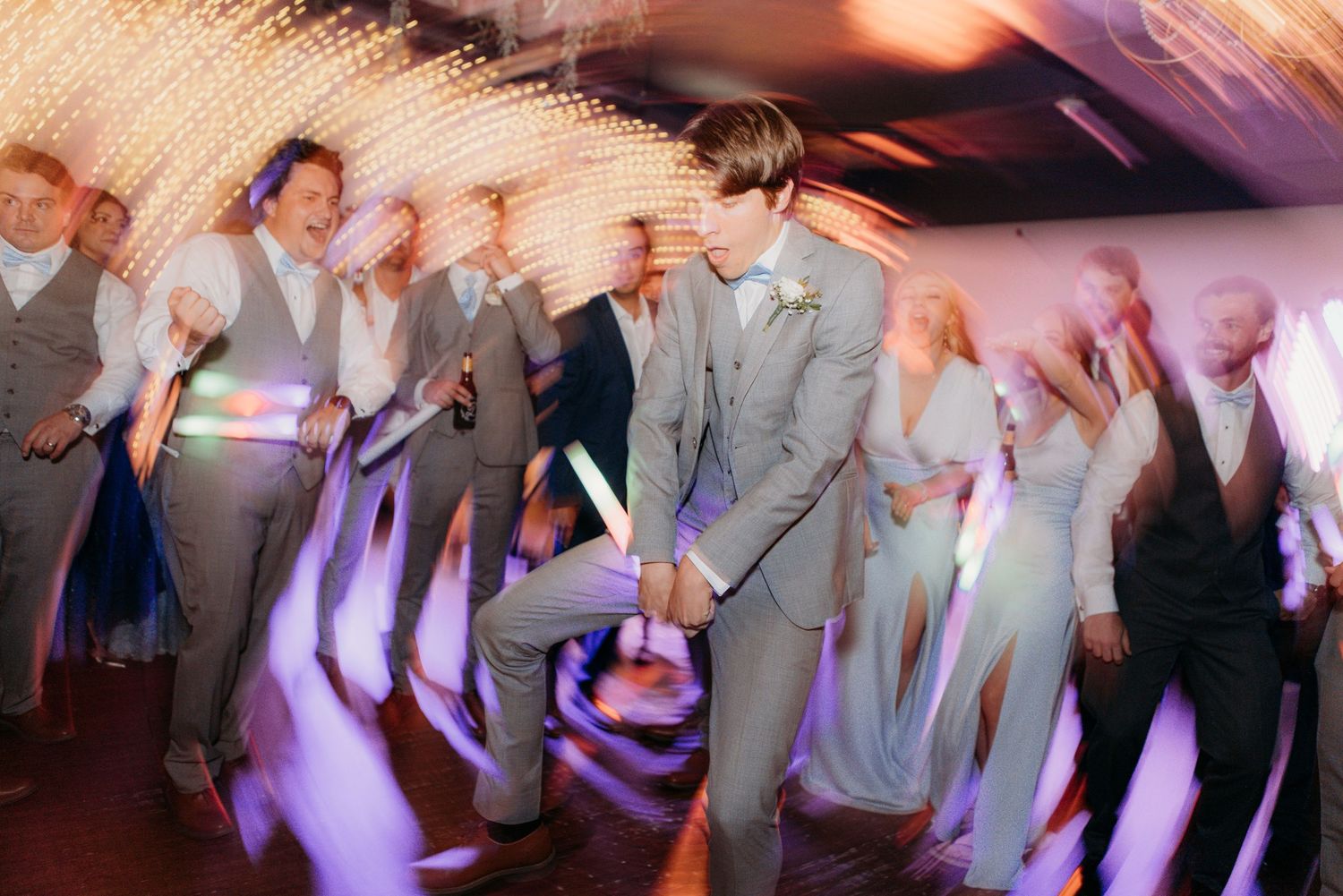 Blurred motion shot of wedding guests dancing energetically at a reception with purple lighting effects.