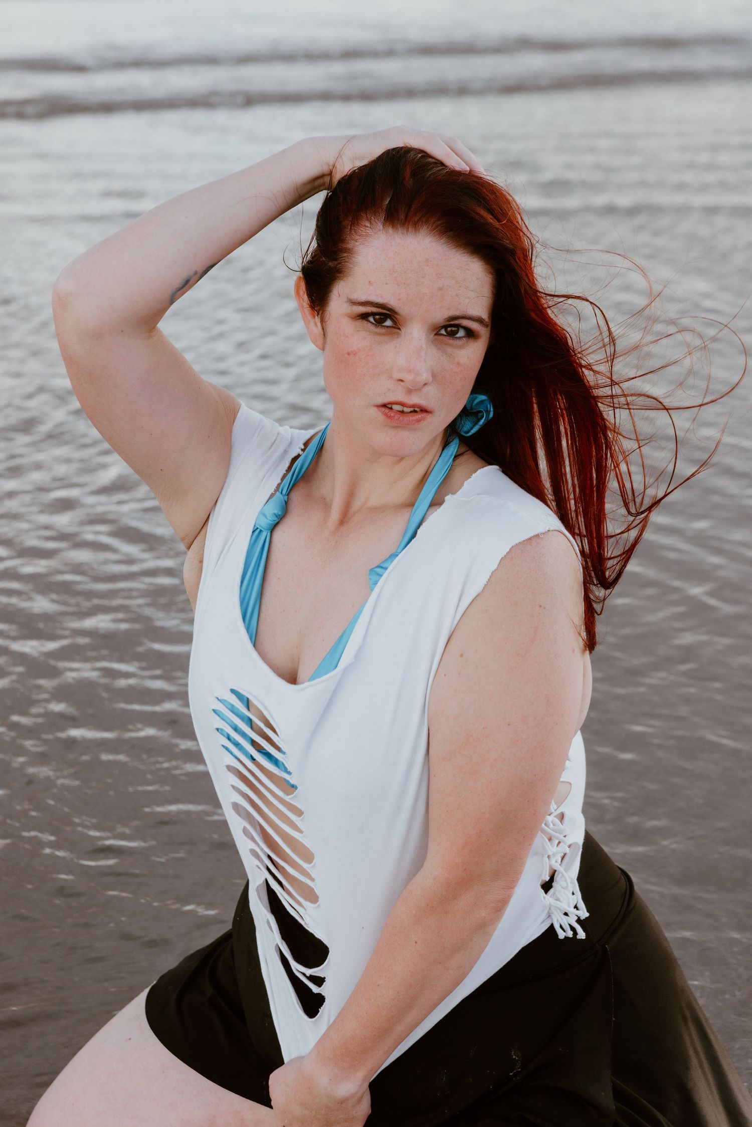 Portrait of someone in a white sleeveless top with blue straps posing near water during sunset with windswept hair.