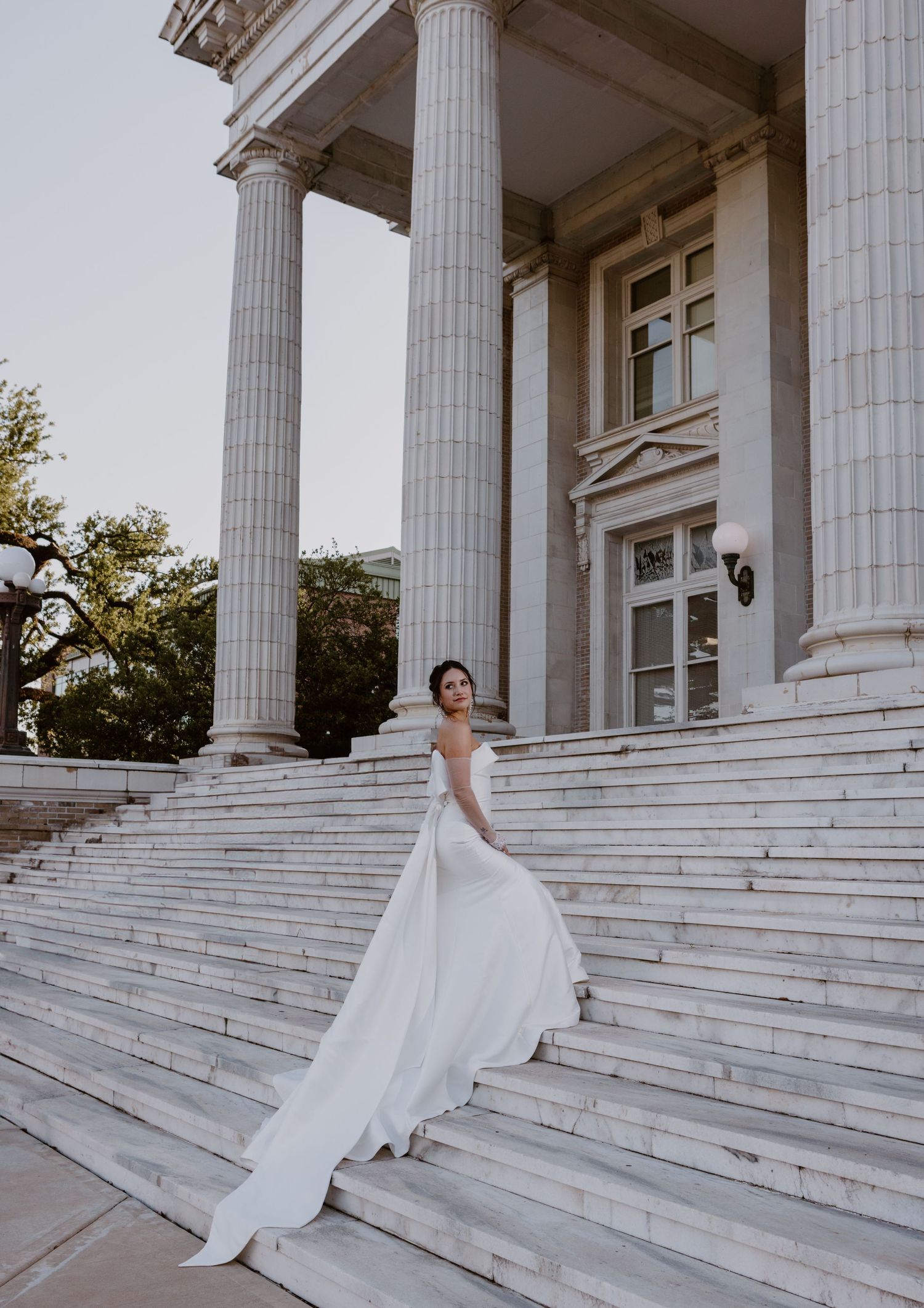 Elegant white wedding dress flowing down grand marble steps of a neoclassical building with tall columns at sunset.