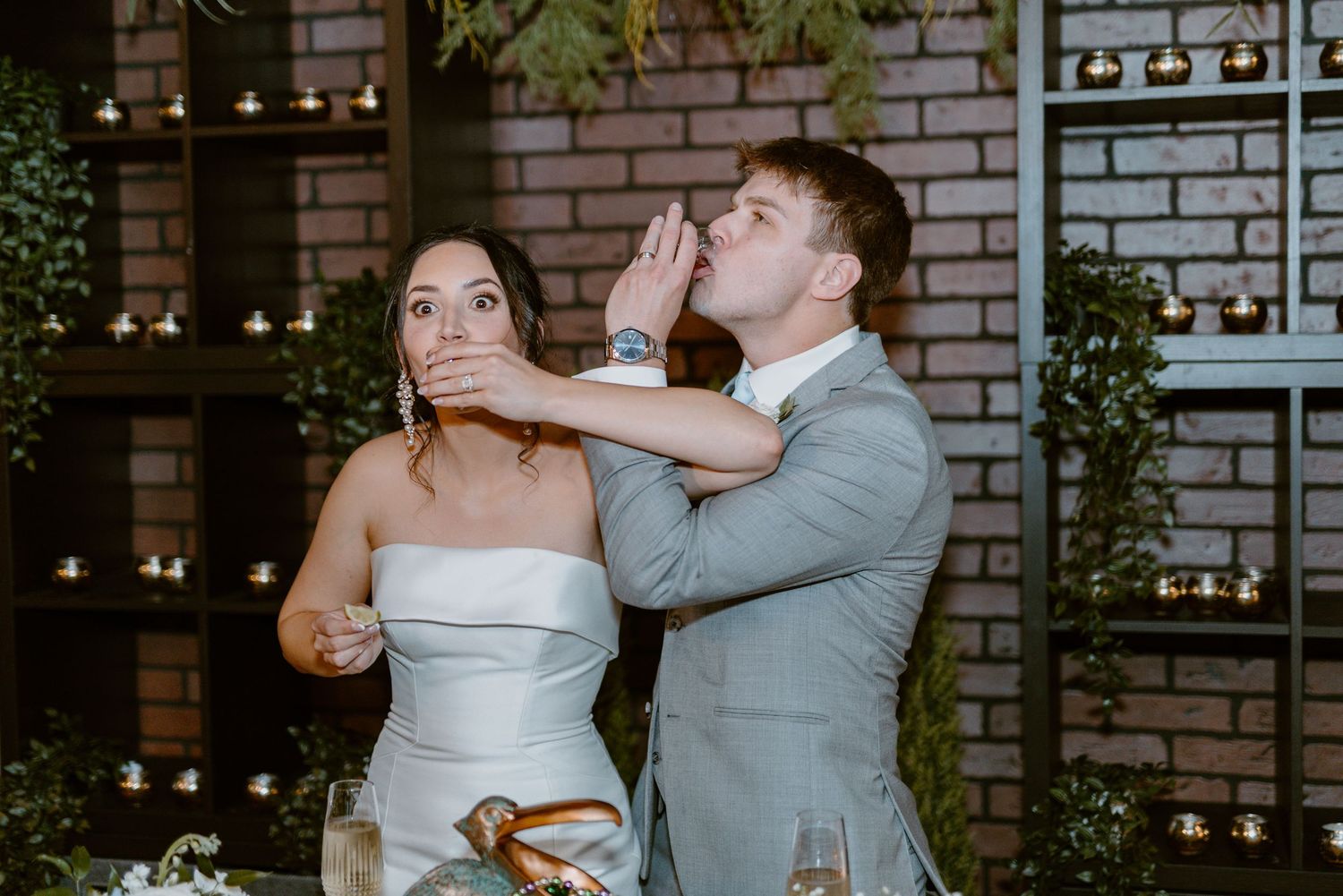 Couple shares playful moment while eating cake at their wedding reception against brick wall with greenery.