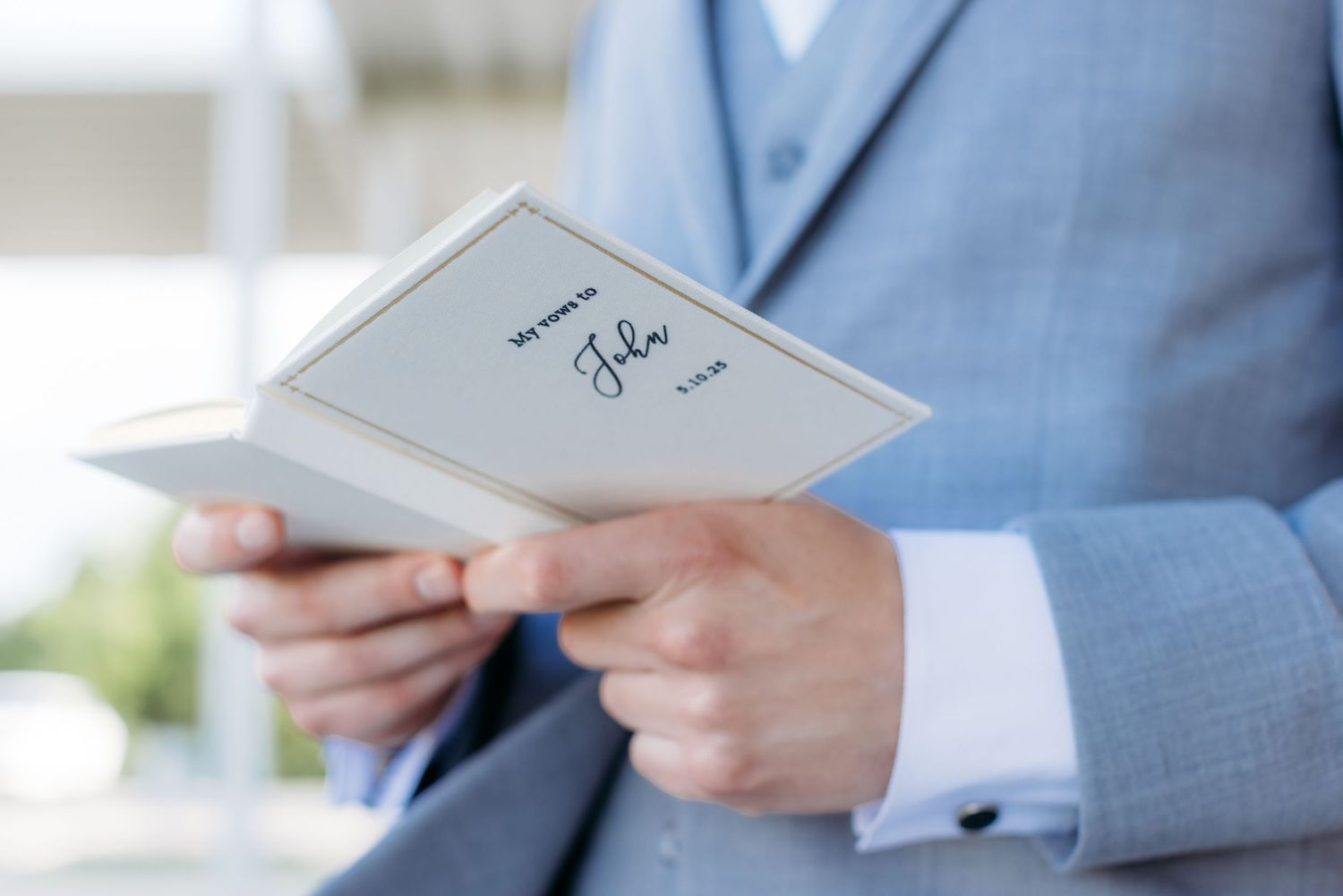 A close-up view of hands holding a light gray linen wedding vow book against a light blue suit.