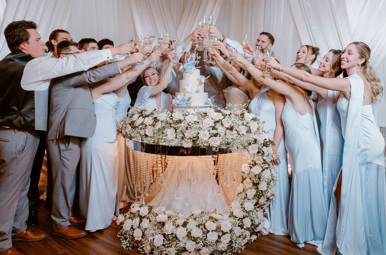 Wedding party toasts and cheers around an elegantly decorated white floral cake display at a reception.
