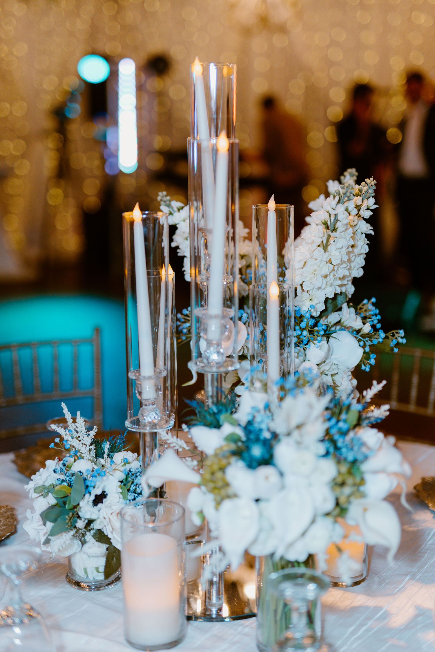 An elegant wedding reception table centerpiece with tall glass candle holders and white floral arrangements in blue lighting.