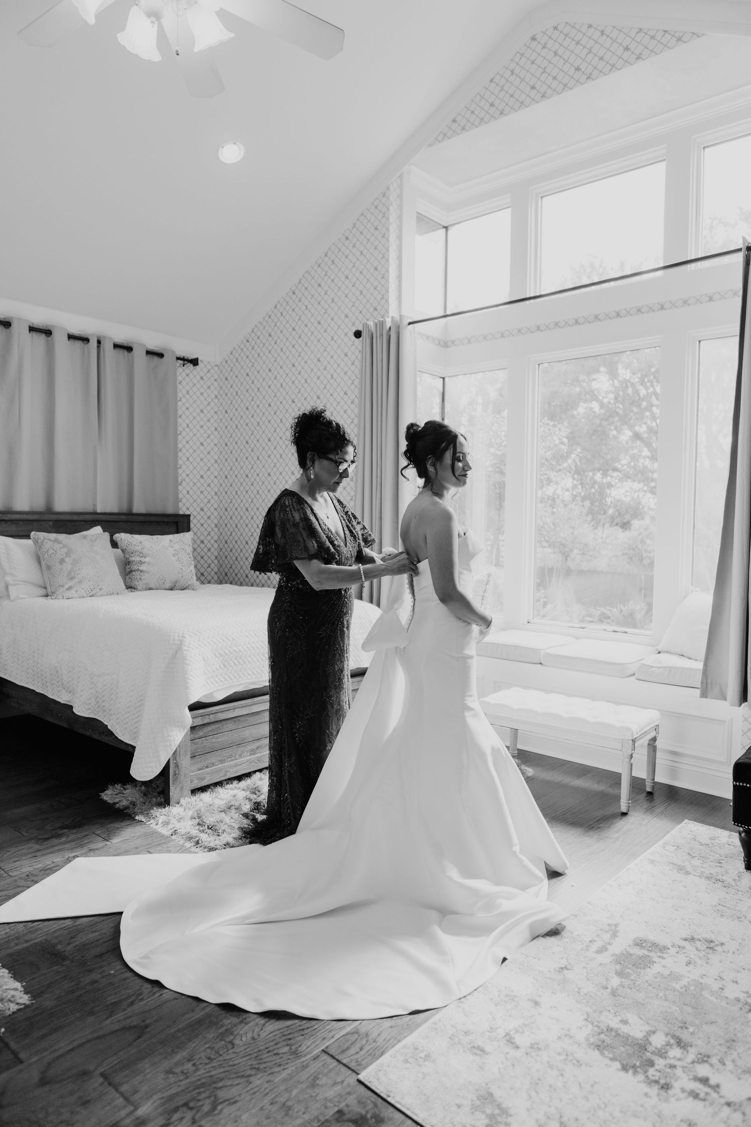 Black and white photo showing a wedding dress fitting in a bright bedroom with a large window and white bedding.