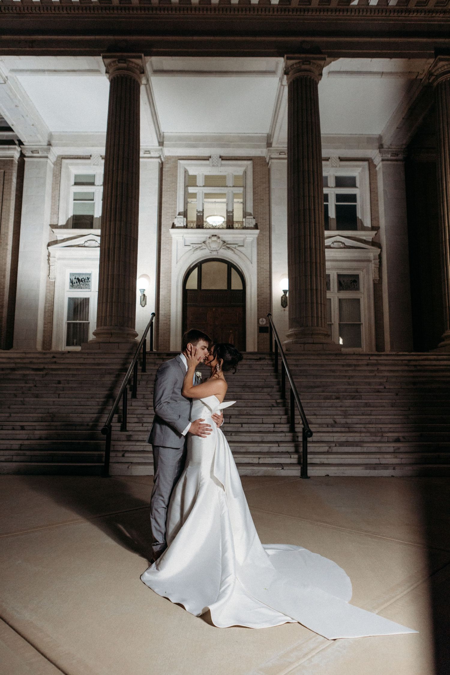 Couple shares romantic moment on grand marble steps of historic building with dramatic columns and arched entrance at night.