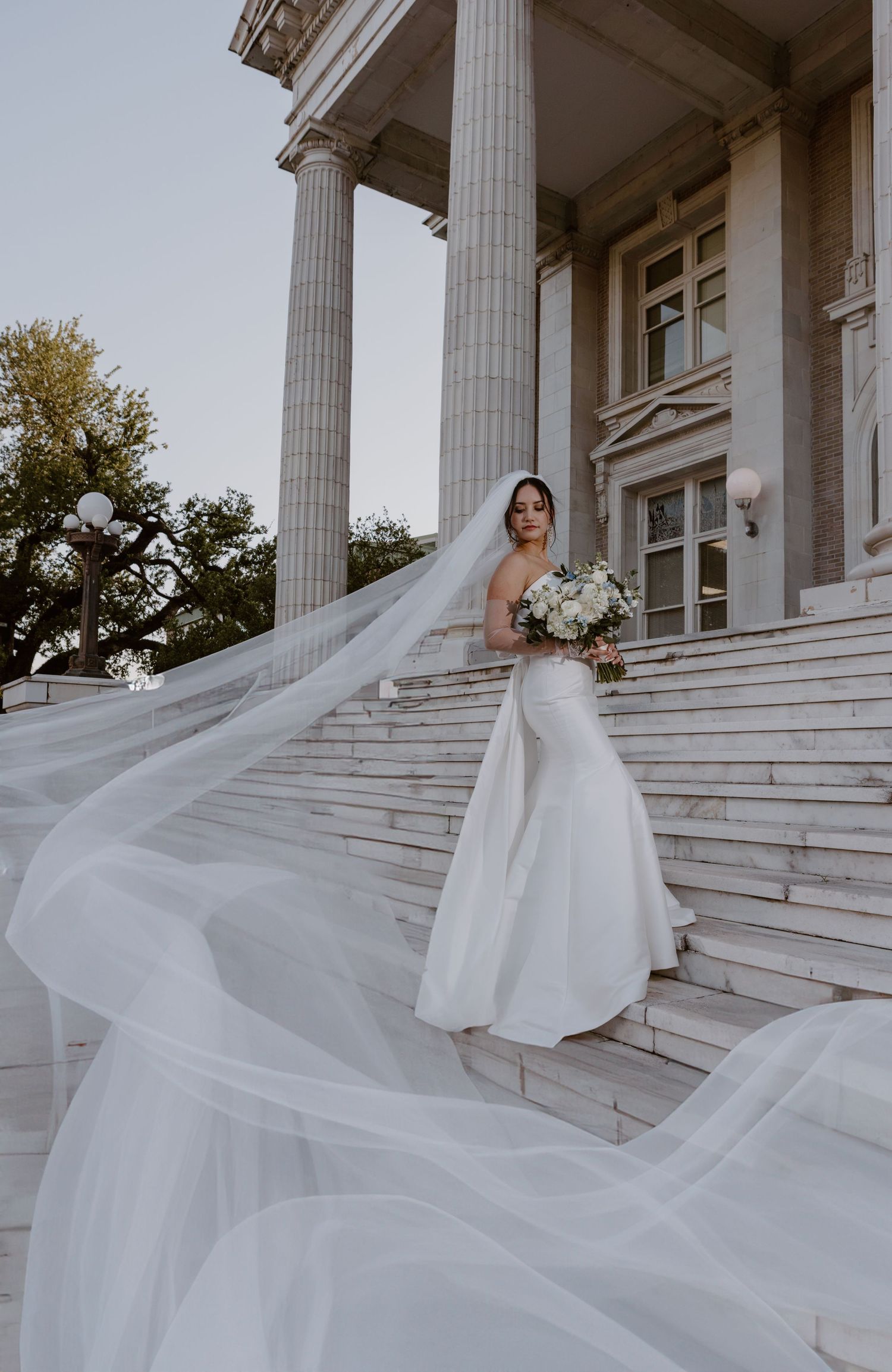 A bride in a flowing white gown stands on marble steps before grand columns as her long veil dramatically billows in the wind.