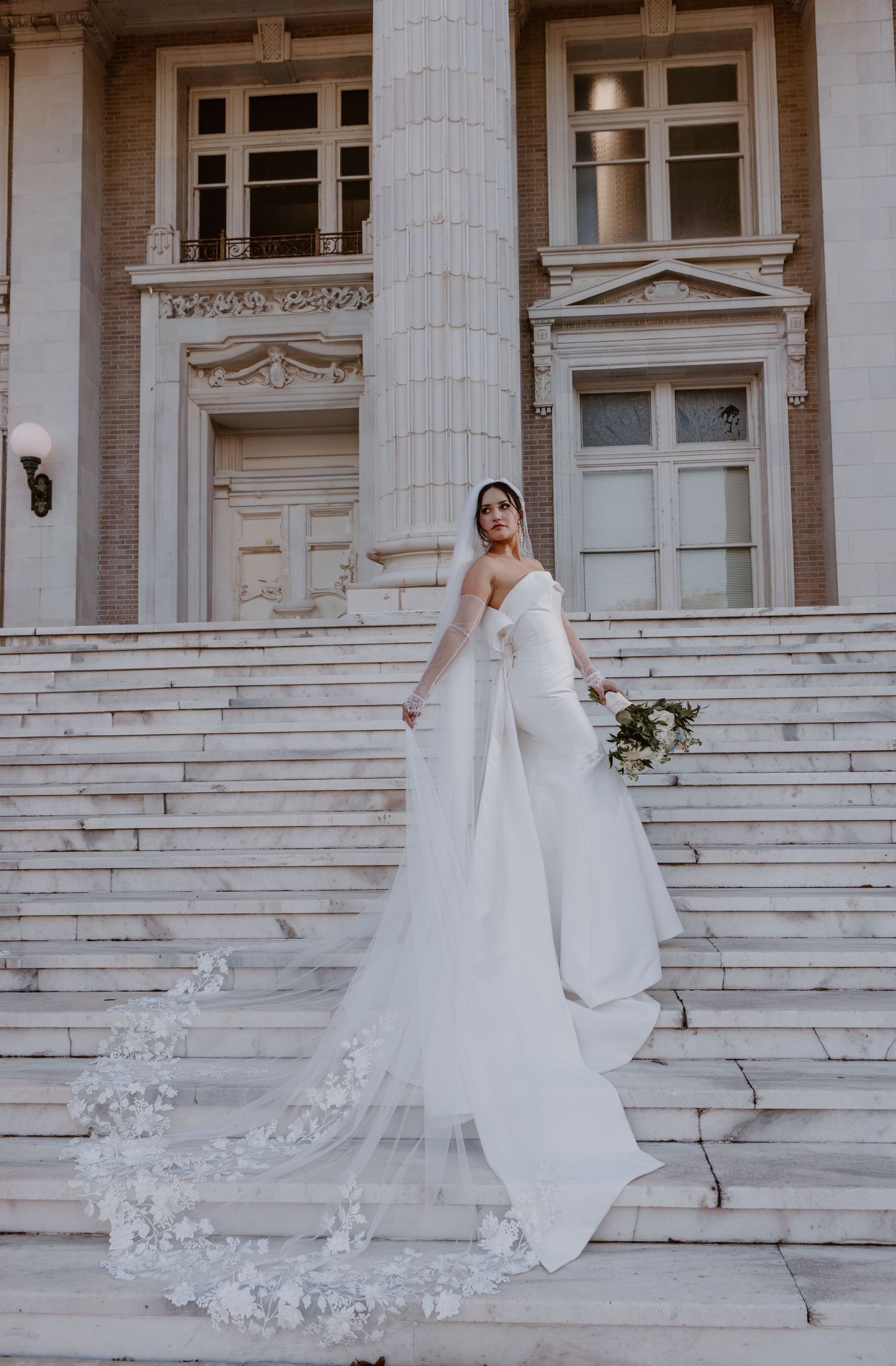 A bride in an elegant white gown and long veil stands on grand marble steps of a historic building with ornate architecture.