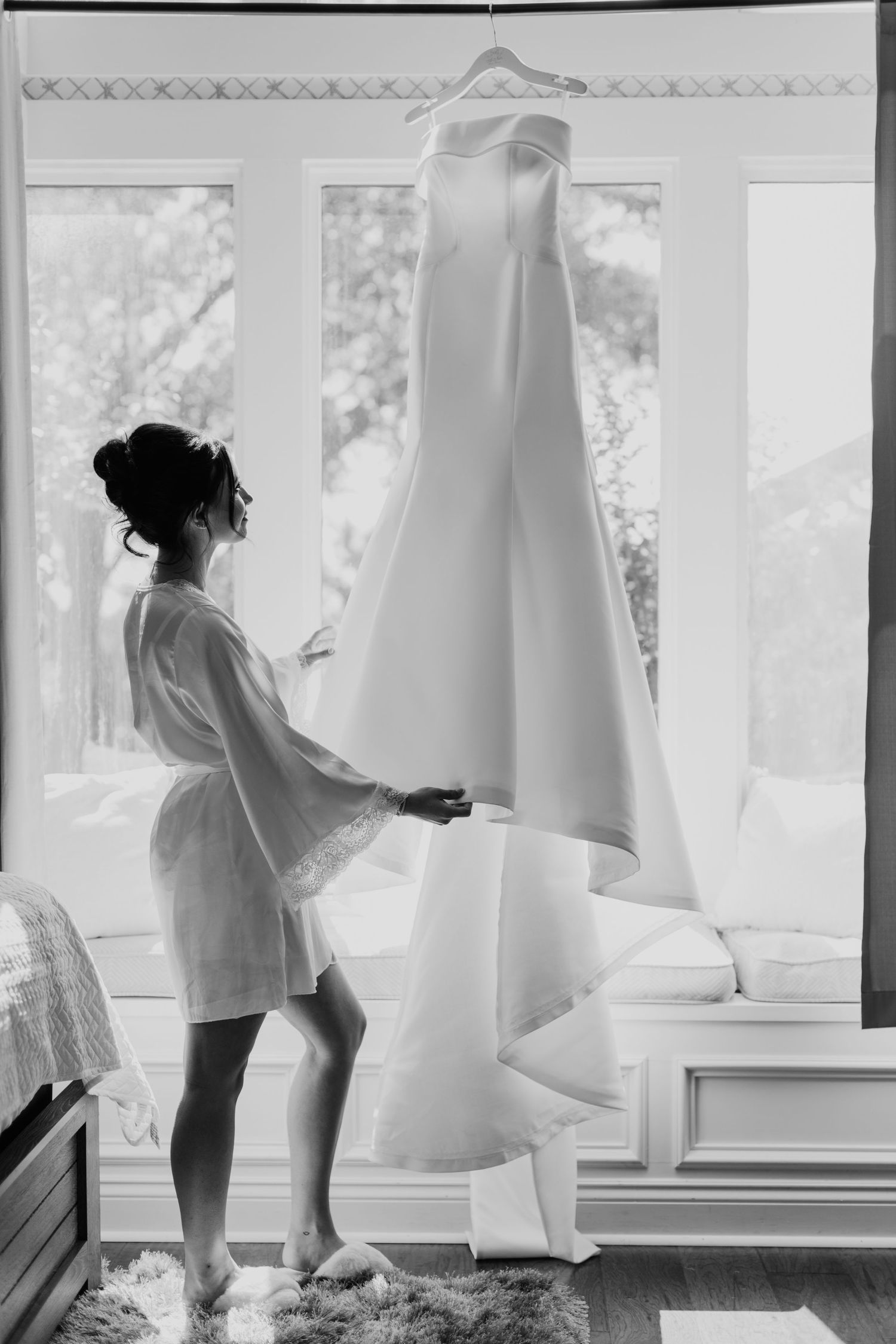 Black and white photo showing elegant bridal dress hanging by window as person in robe admires gown on wedding morning.
