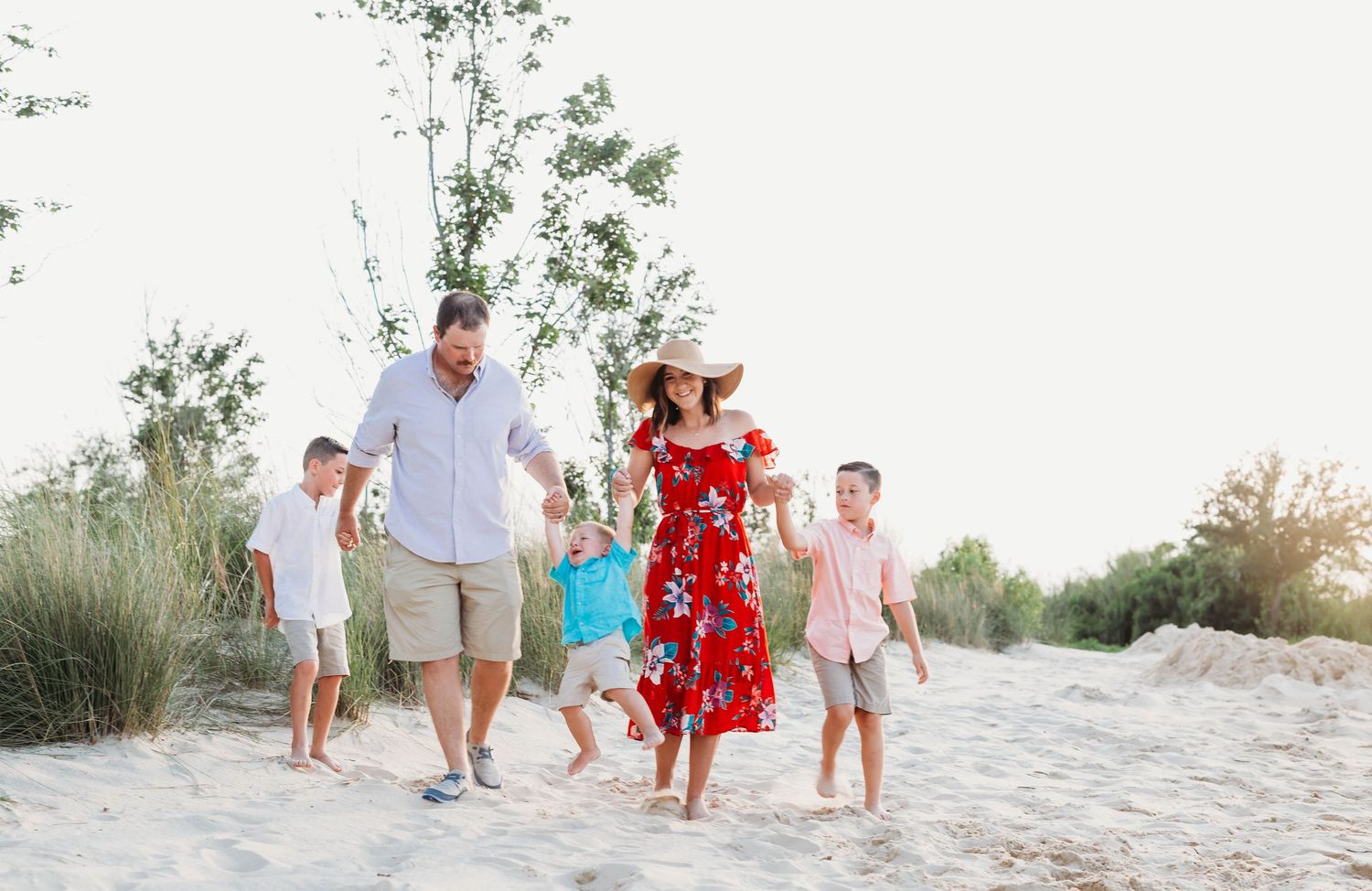 Family of four walking together on sandy beach path during sunset while wearing casual summer attire.