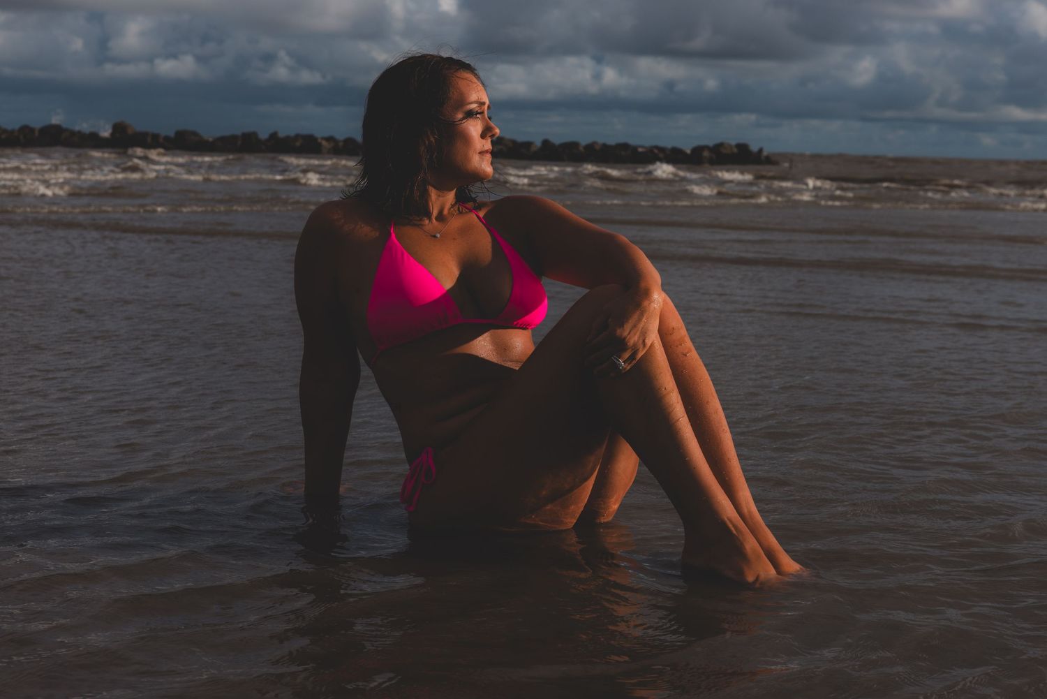 A person in a swimsuit sitting at the edge of the ocean as waves crash around during sunset creating a silhouette effect.
