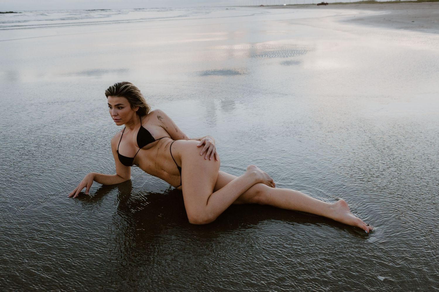 Woman in swimwear poses on wet sand at the beach during overcast day.