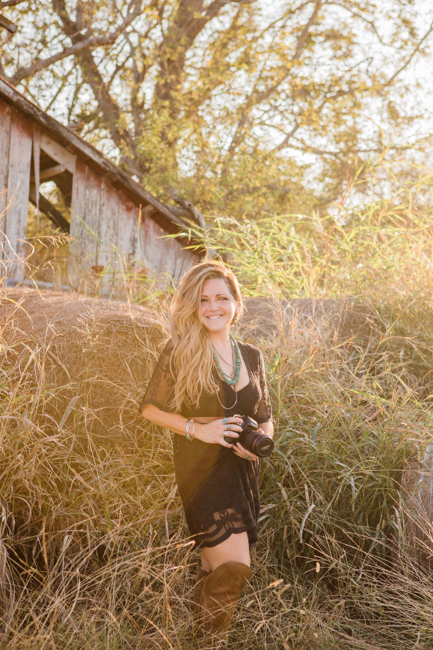 A photographer in a black dress holds a camera in a golden sunlit field of tall grass.