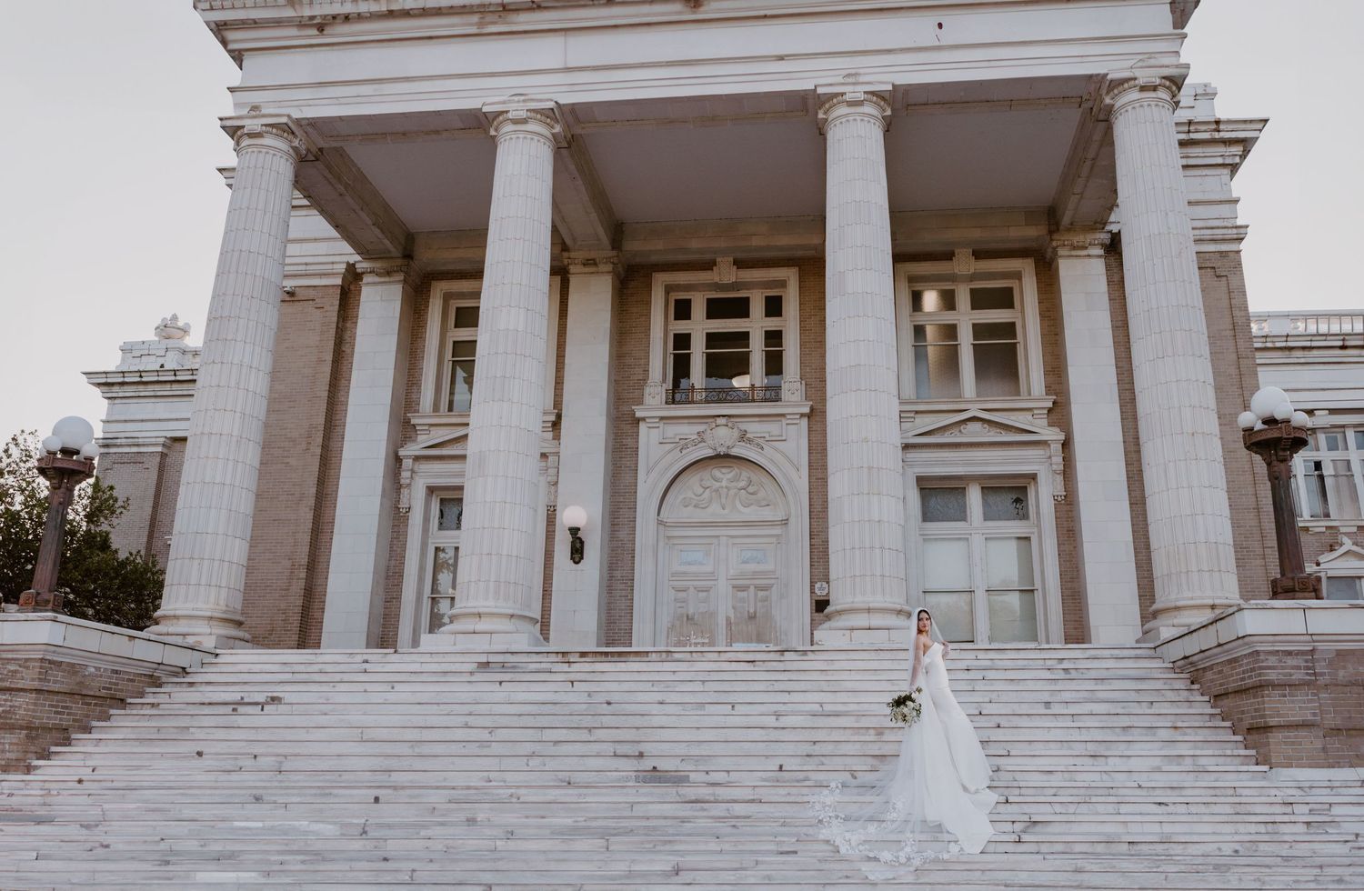 A bride in a white wedding dress stands on the grand steps of a neoclassical building with tall columns.