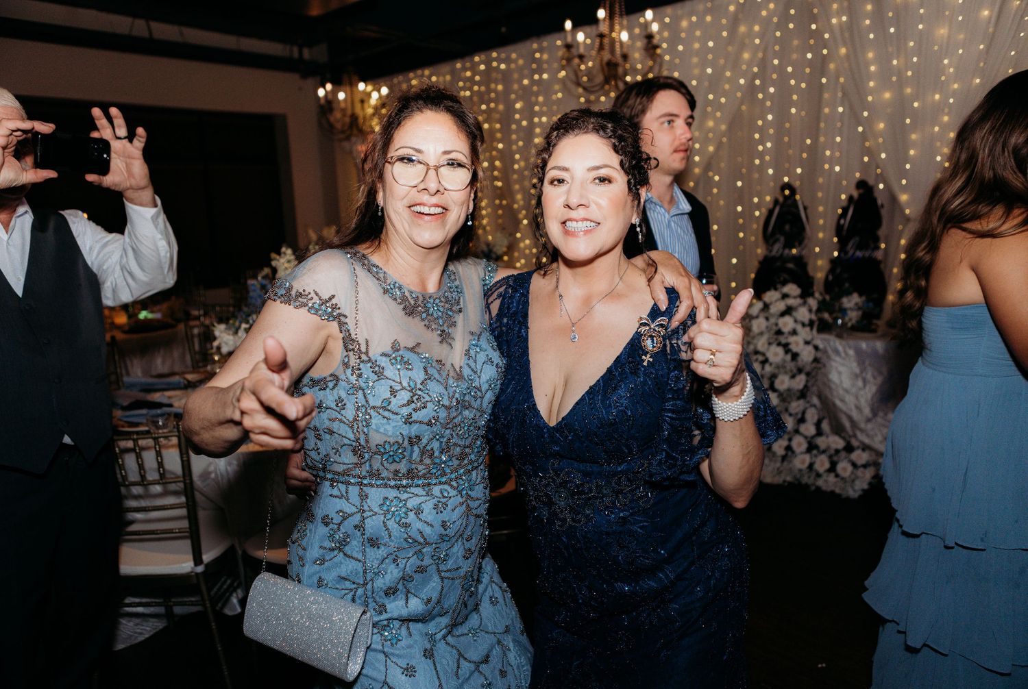 Party guests in blue dresses dance and celebrate at an event with twinkle lights in the background.