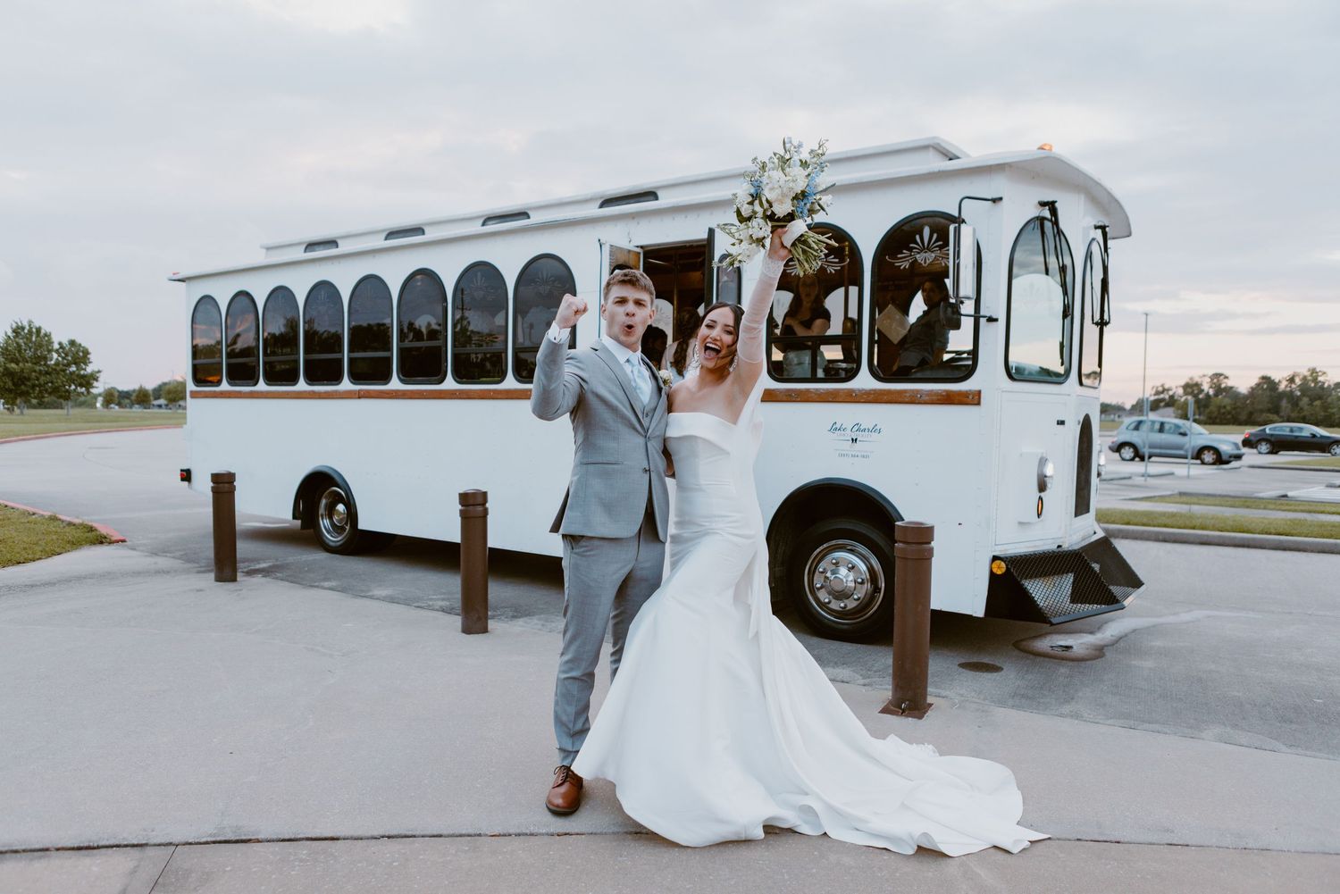 A newlywed couple celebrates in front of a white vintage trolley bus at an outdoor venue.