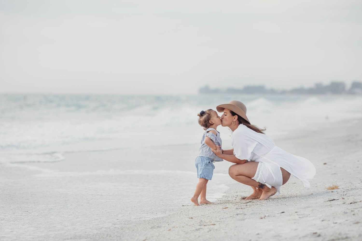 Person in white dress crouches next to small child on misty beach at sunset while waves roll in behind them.