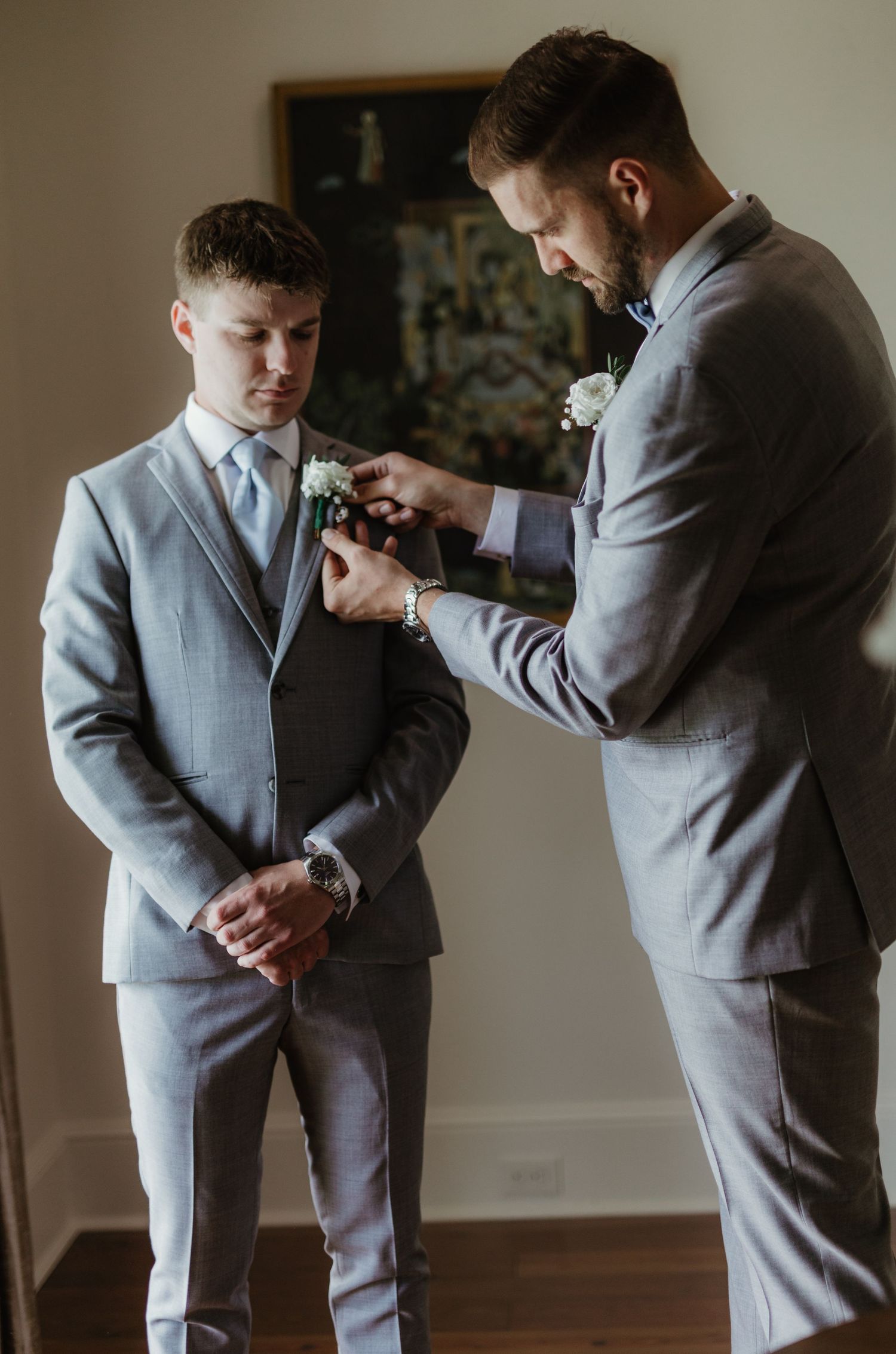 Two people in gray suits preparing for a formal event as one helps adjust the other's boutonniere.