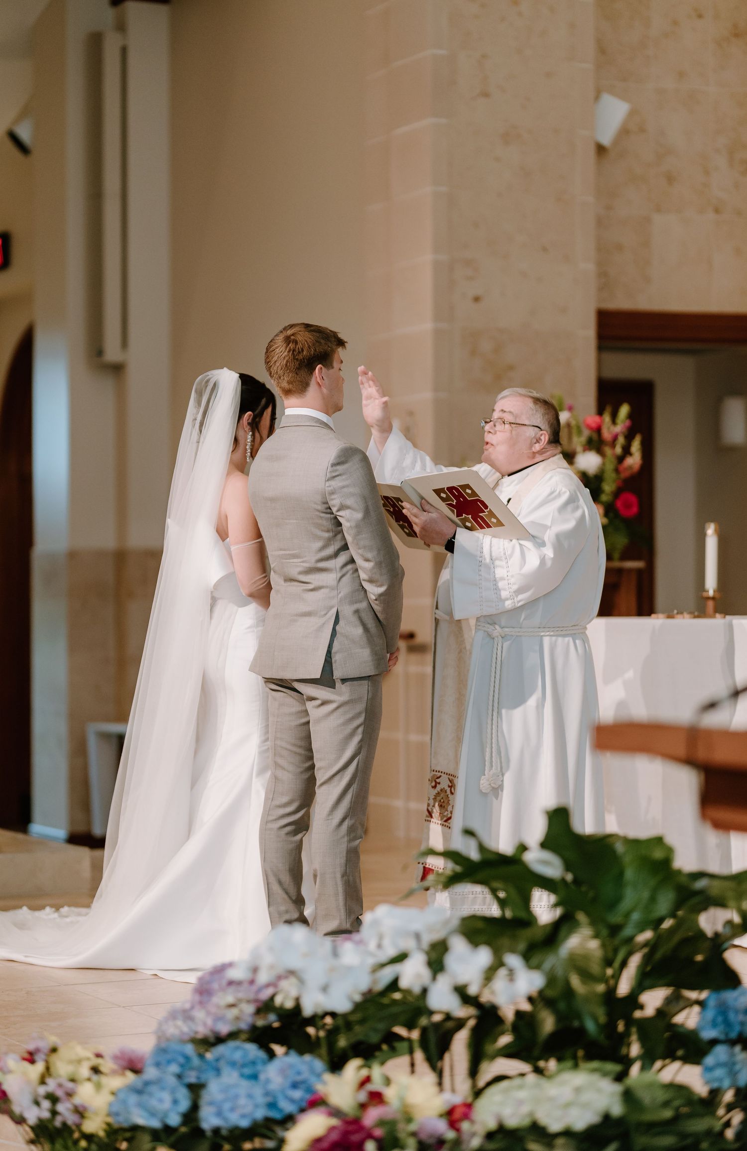 A Catholic priest presides over a wedding ceremony with a bride and groom at an altar surrounded by flowers.