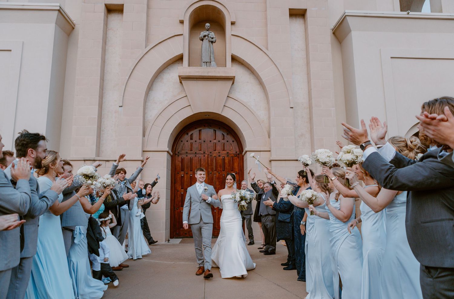 Wedding party wearing light blue dresses forms pathway outside church entrance during celebration ceremony.