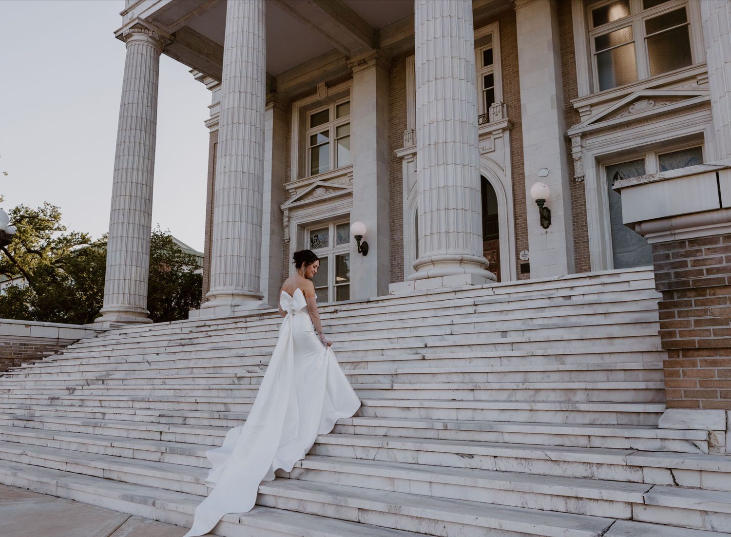 A person in a flowing white wedding gown stands on marble steps of a grand neoclassical building with tall columns.
