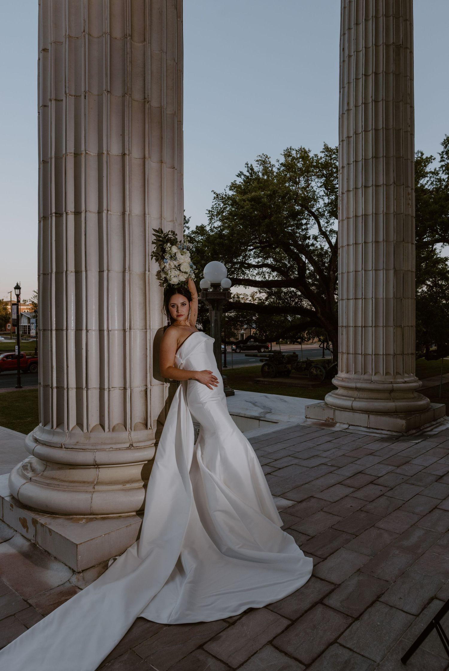 An elegant wedding dress with a flowing train is photographed between classical stone columns at dusk.