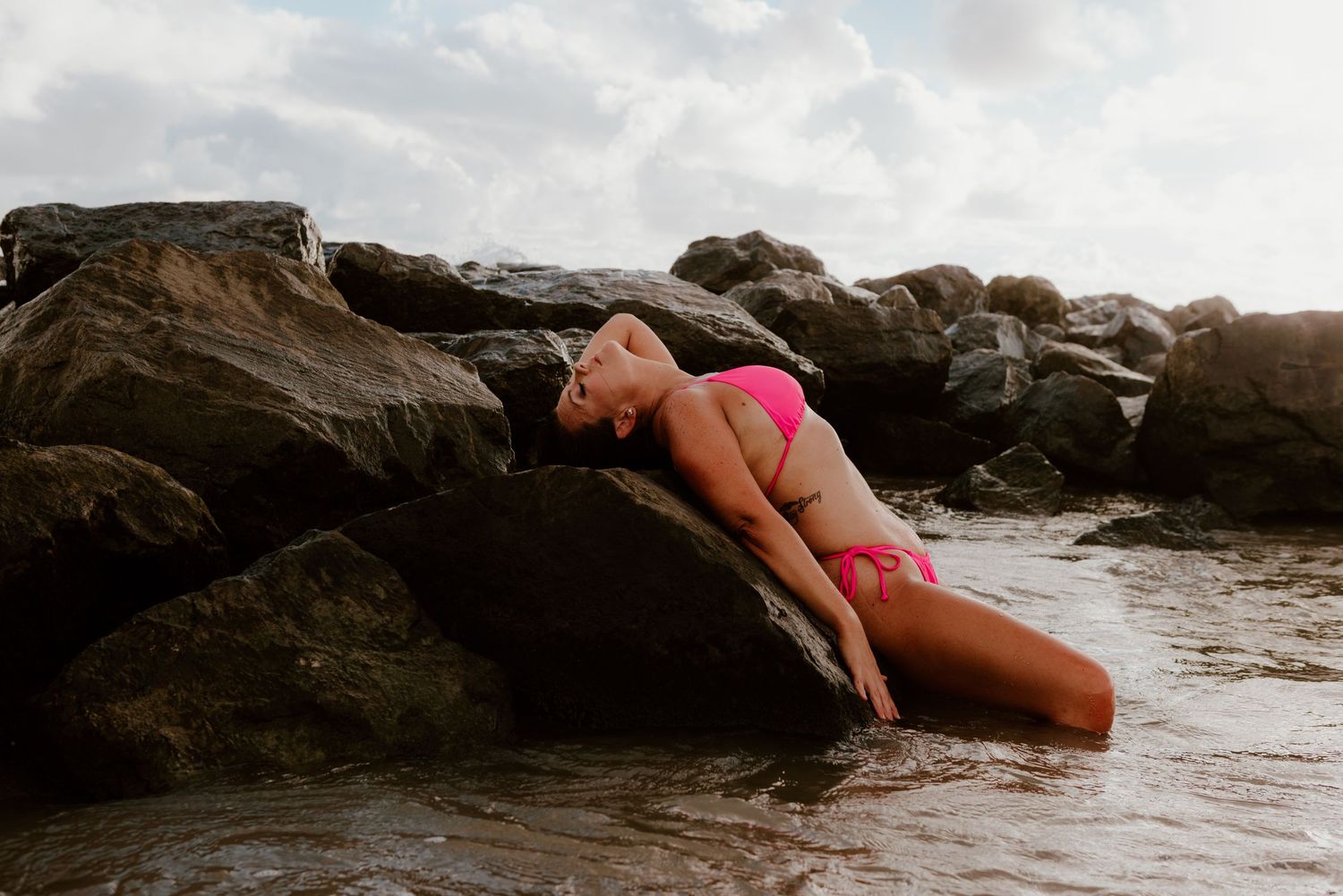 Figure reclining on coastal rocks near ocean at sunset wearing swimwear and basking in golden light.