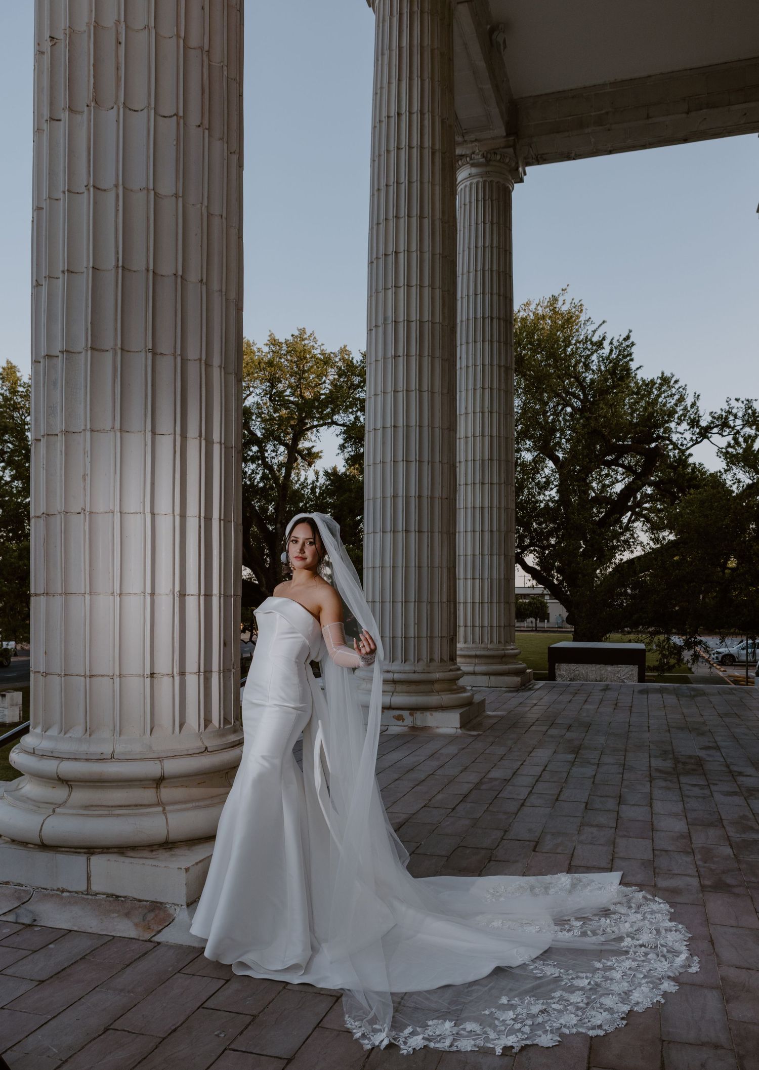 An elegant wedding dress with a flowing lace train is showcased between grand classical columns at dusk.