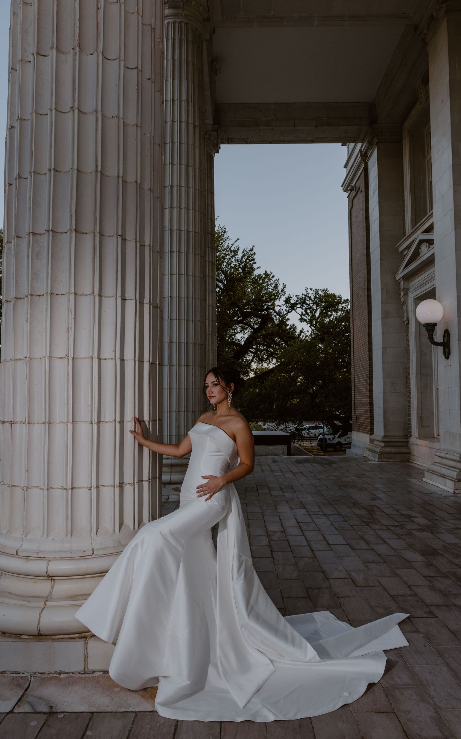 Elegant bridal portrait showing a strapless white wedding gown with flowing train against classical stone columns at sunset.