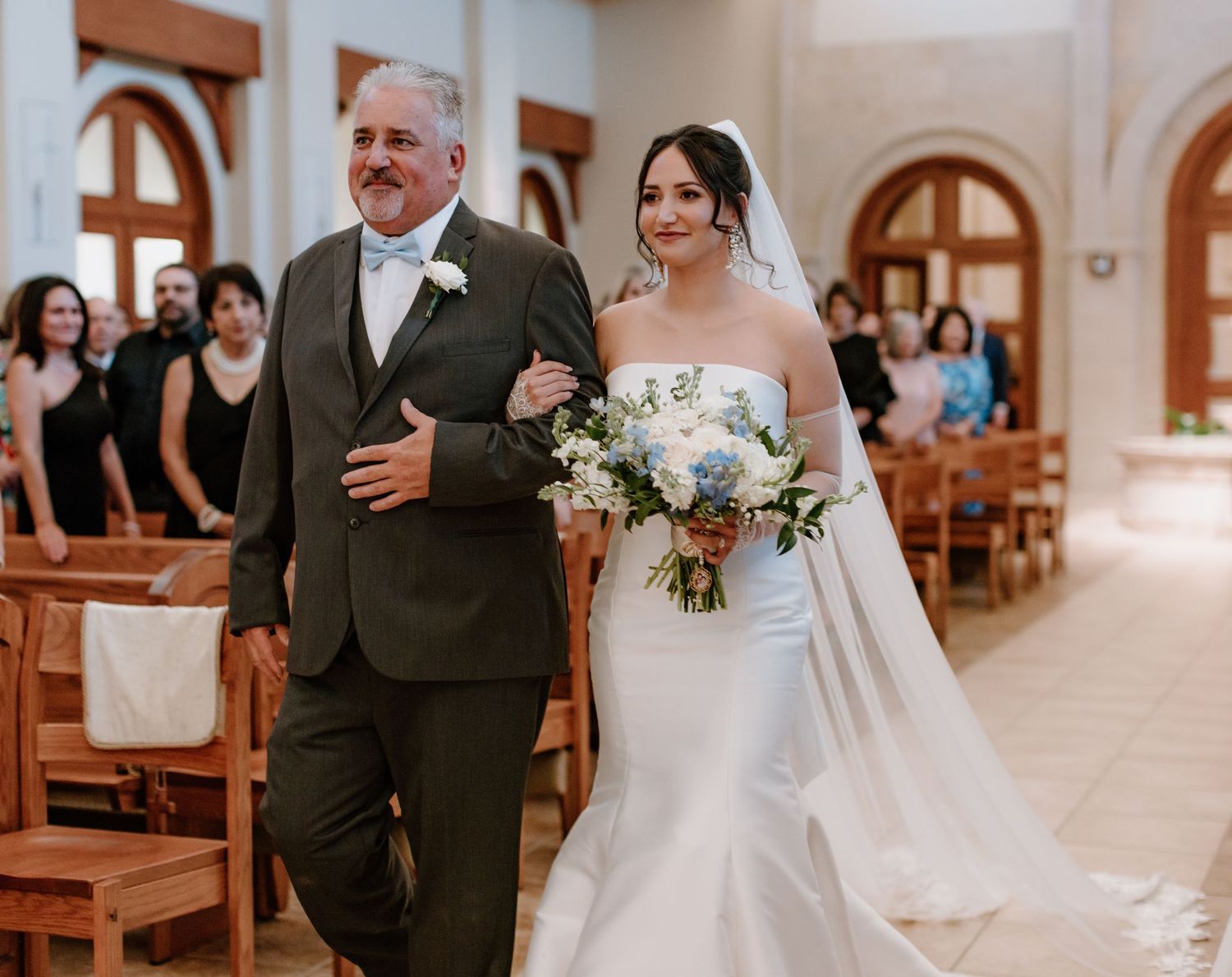 A bride in a white lace gown walks down the church aisle with her escort amid wooden pews and arched windows.