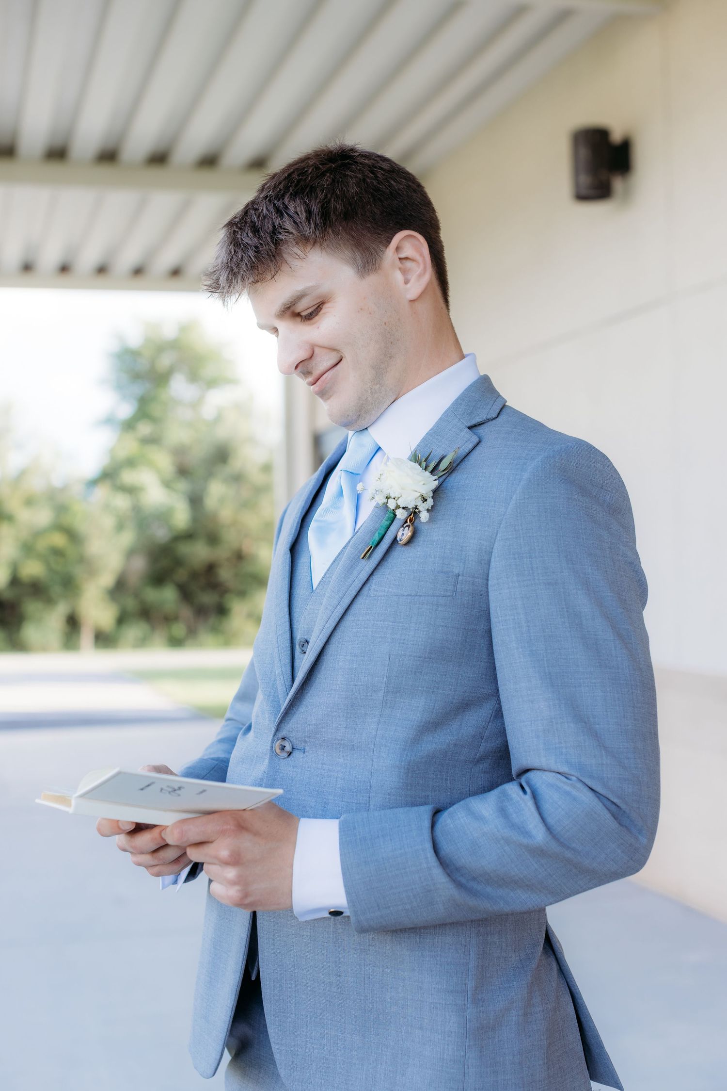A person wearing a light blue suit and boutonniere reads from papers while smiling in an outdoor covered area.
