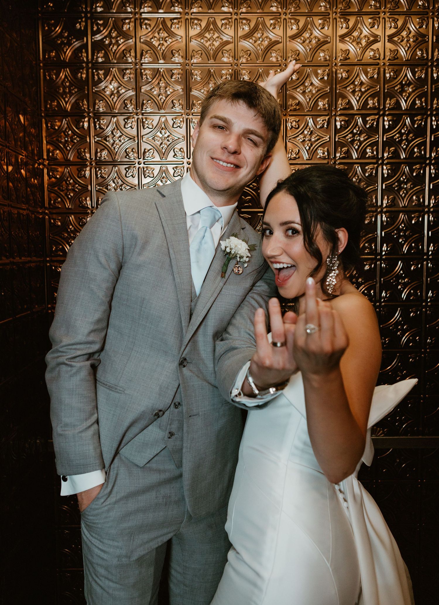 Happy newlyweds show off ring and share joyful moment against metallic textured backdrop.