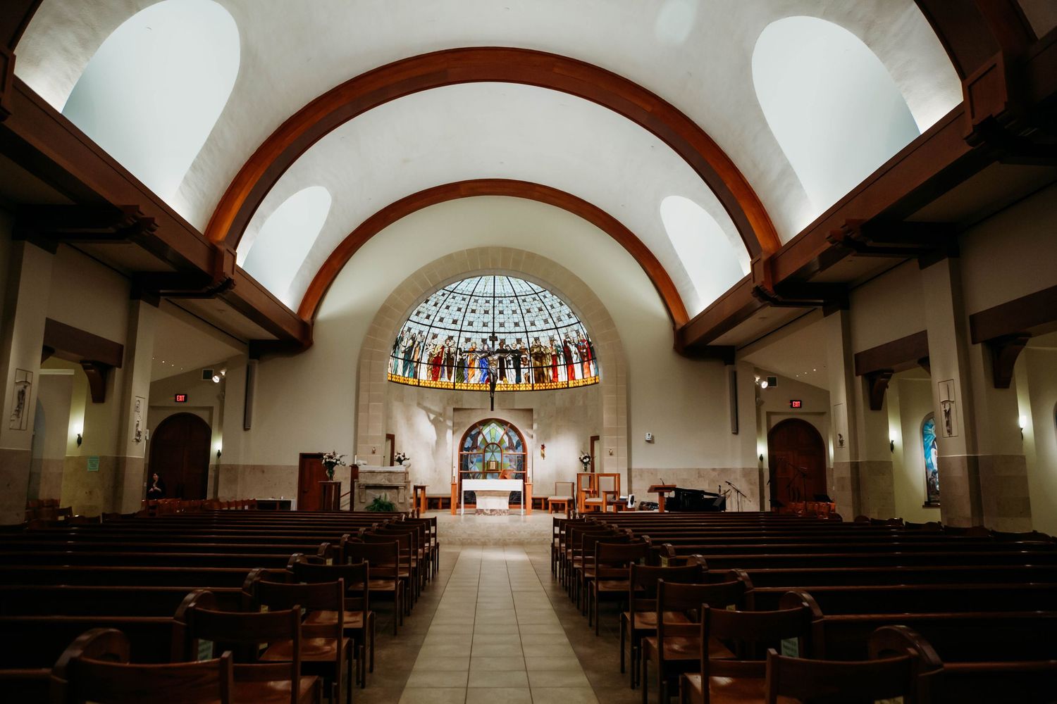 Interior view of a church sanctuary with vaulted arches, wooden pews, and stained glass window at the altar.