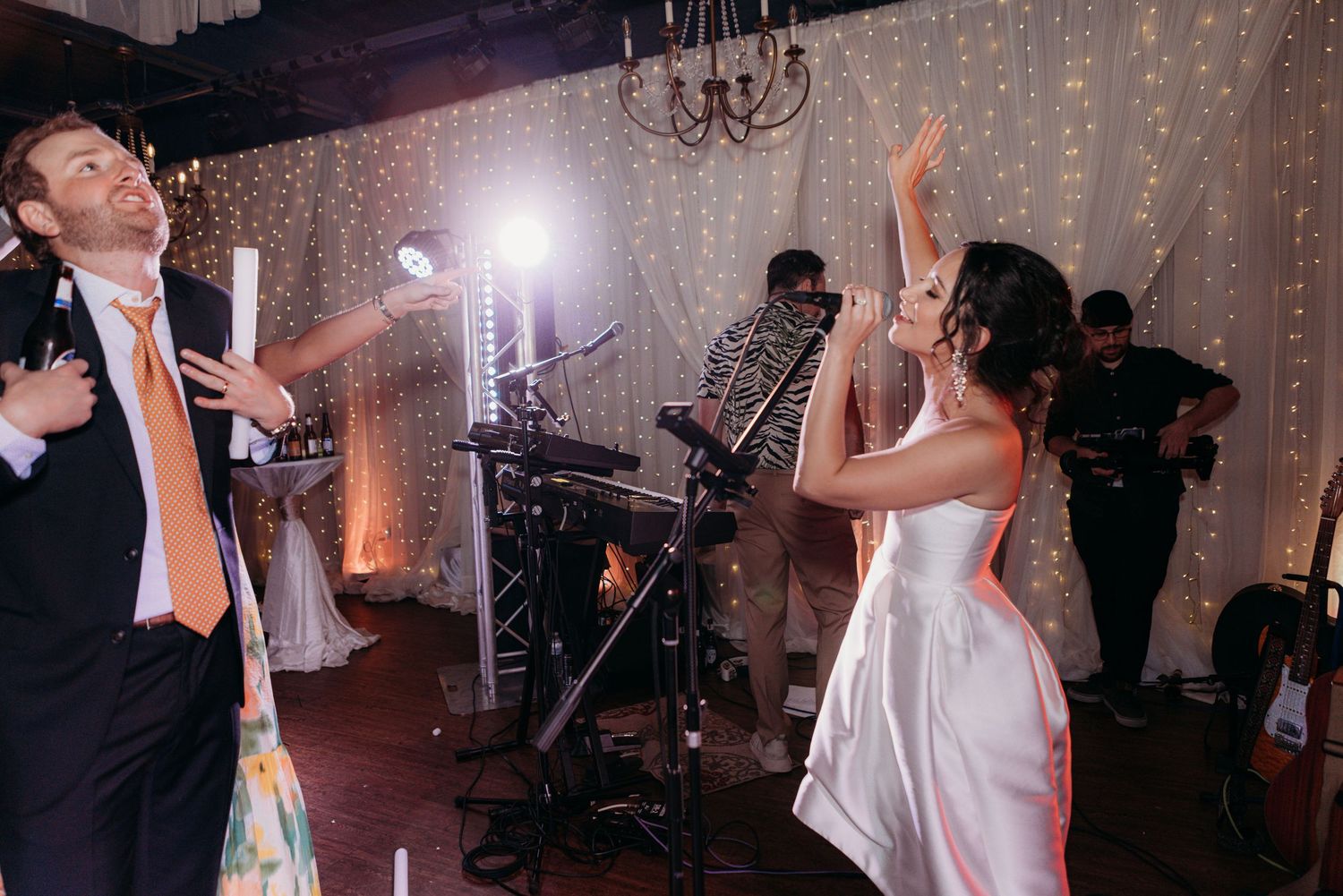 Wedding guests dance and celebrate at an indoor reception with twinkle lights and a chandelier in the background.